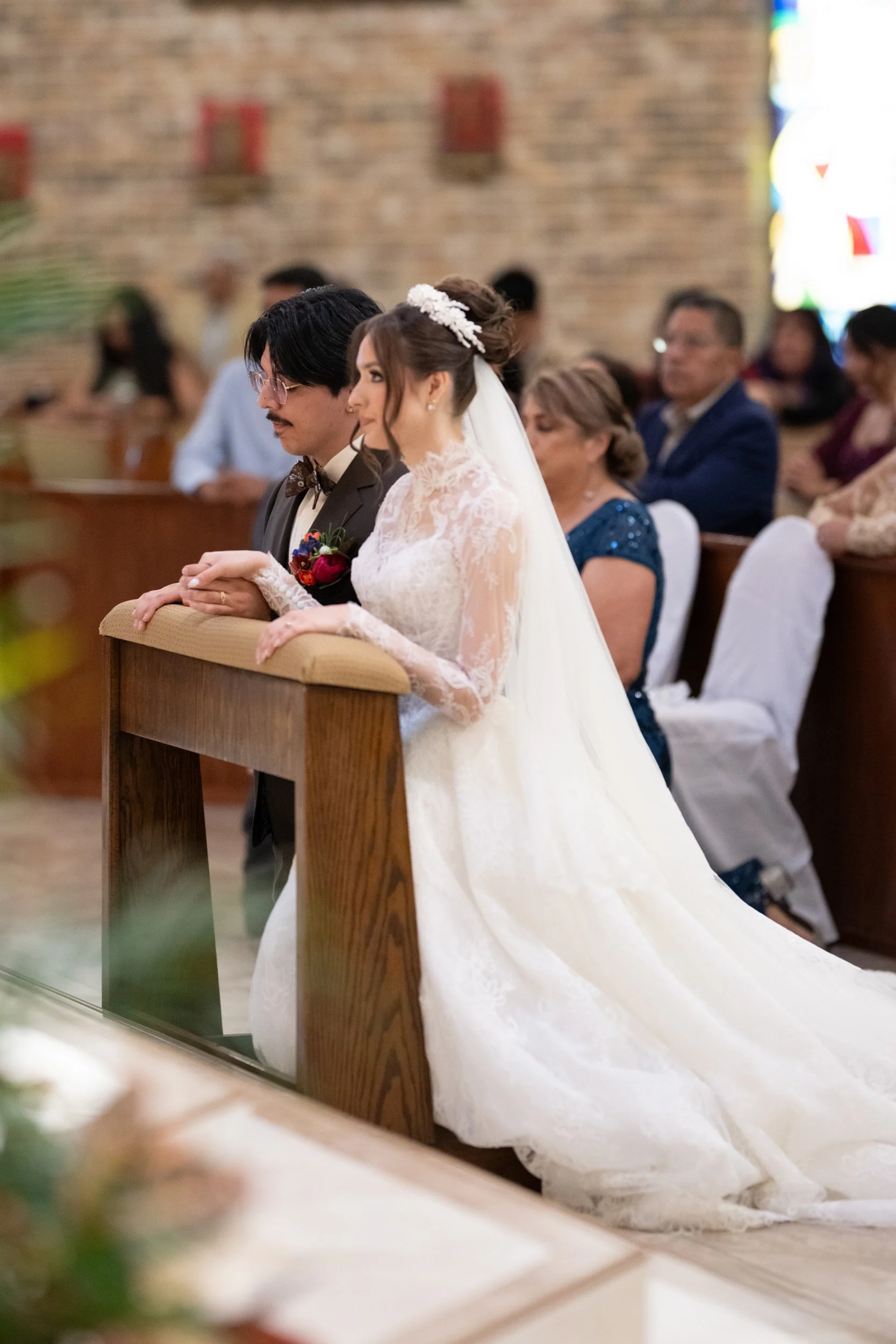 Bride and groom kneeling together in prayer during a traditional church wedding ceremony, captured from the side as guests look on in a reverent, intimate moment.