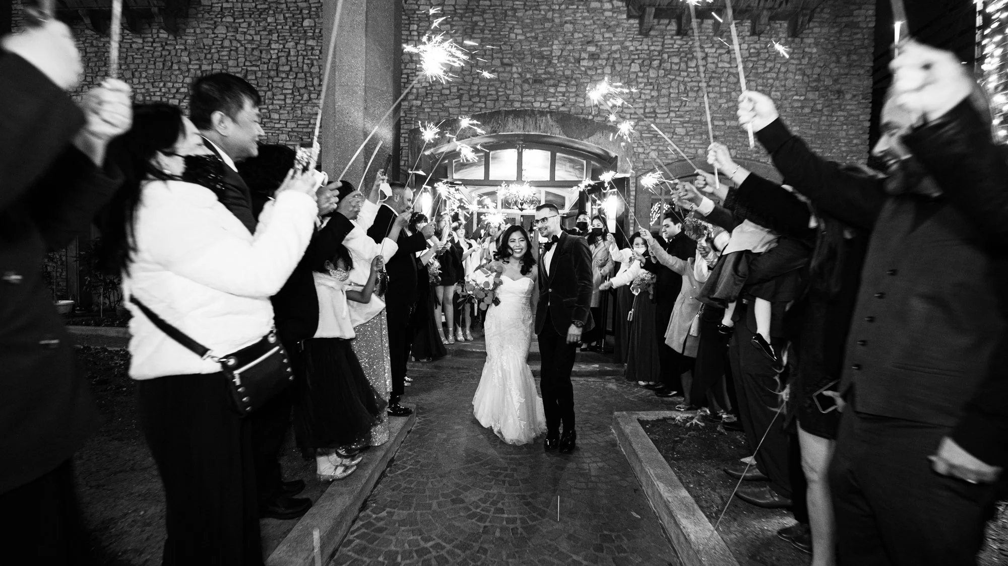 The bride and groom exit their reception through a tunnel of sparklers, surrounded by cheering friends and family. The glowing sparks and stone architecture create a dramatic, cinematic farewell filled with energy and love.