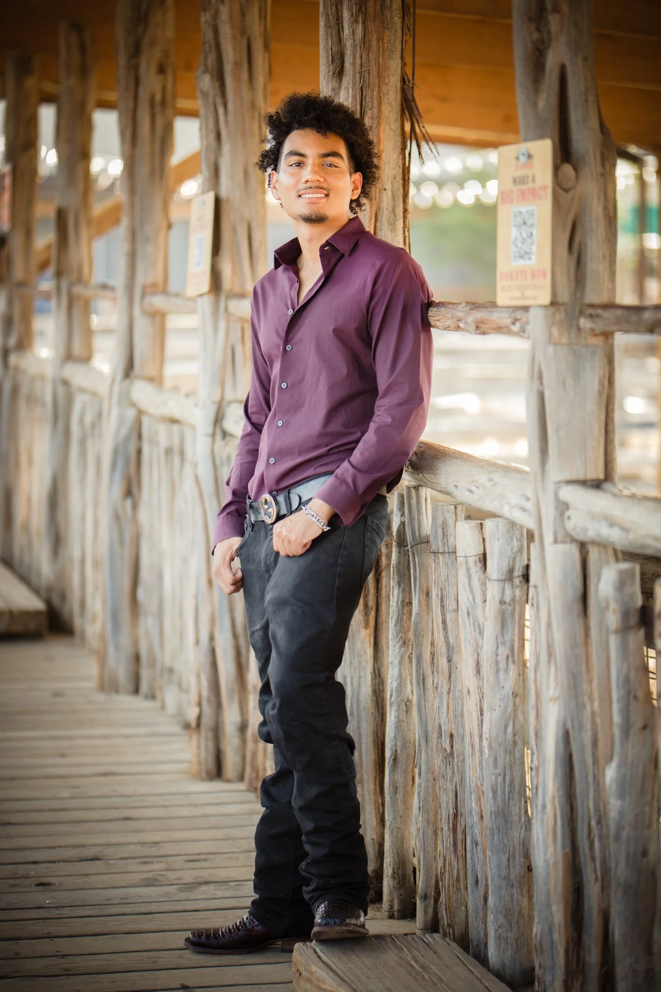 A young man in a maroon button-down shirt and dark jeans poses along a rustic wooden walkway, illuminated by natural light for a relaxed portrait style.