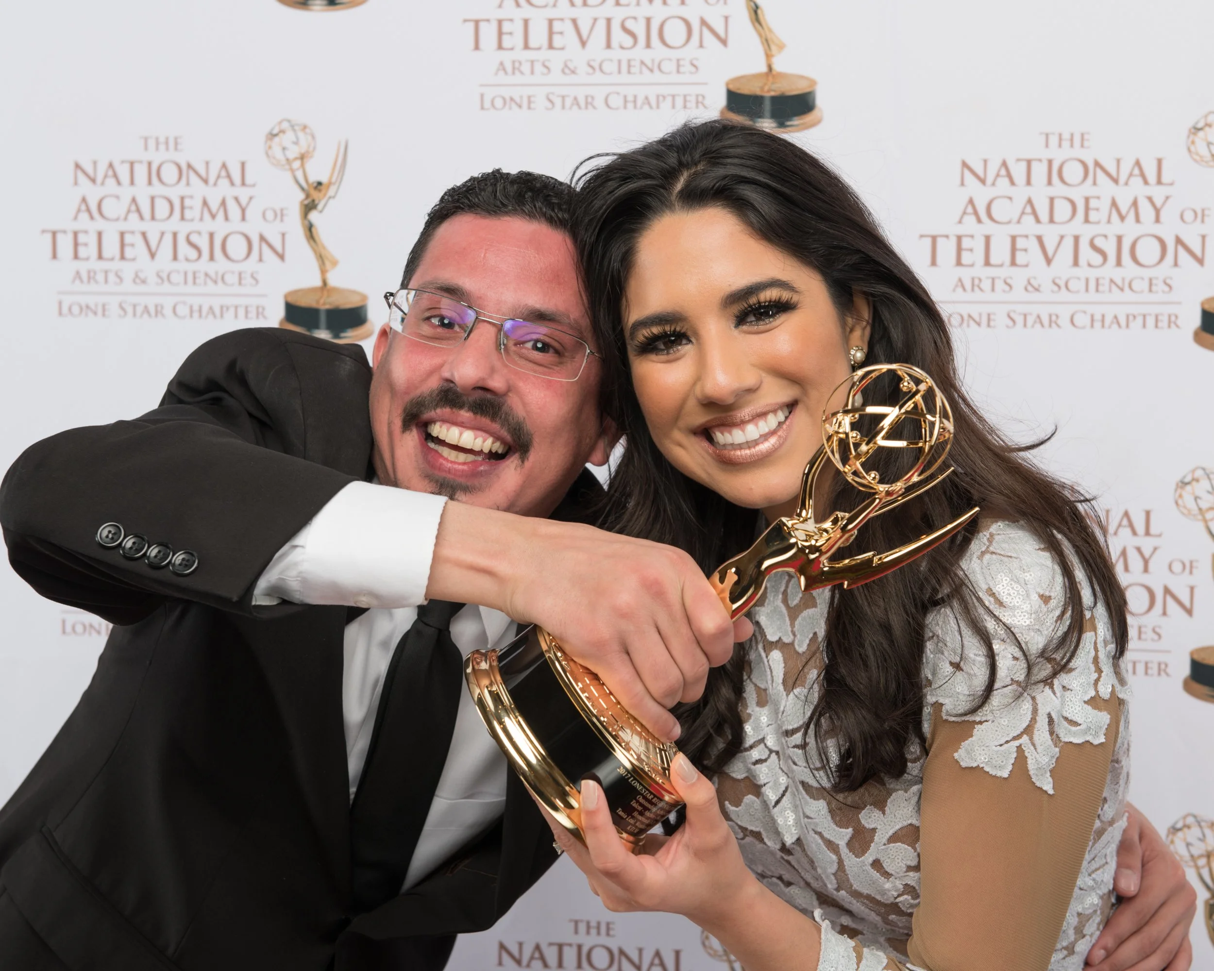 A pair of Emmy Award winners pose closely while holding an Emmy trophy, smiling proudly during a red-carpet portrait session at the National Academy of Television Arts & Sciences event.