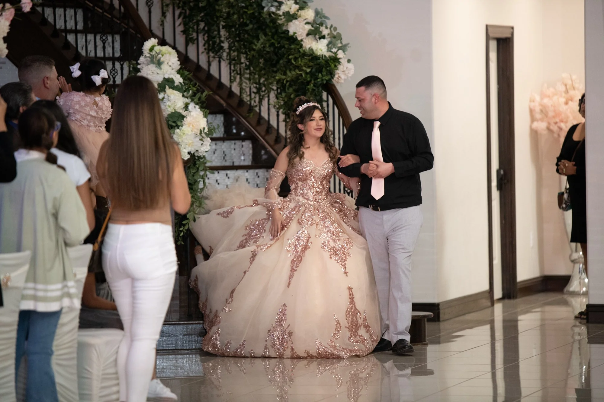 Escorted into the reception space, Katelyn smiles as she makes her way through guests beneath a staircase wrapped in floral greenery. This entrance captures the poise and excitement of stepping fully into her Quinceañera celebration.