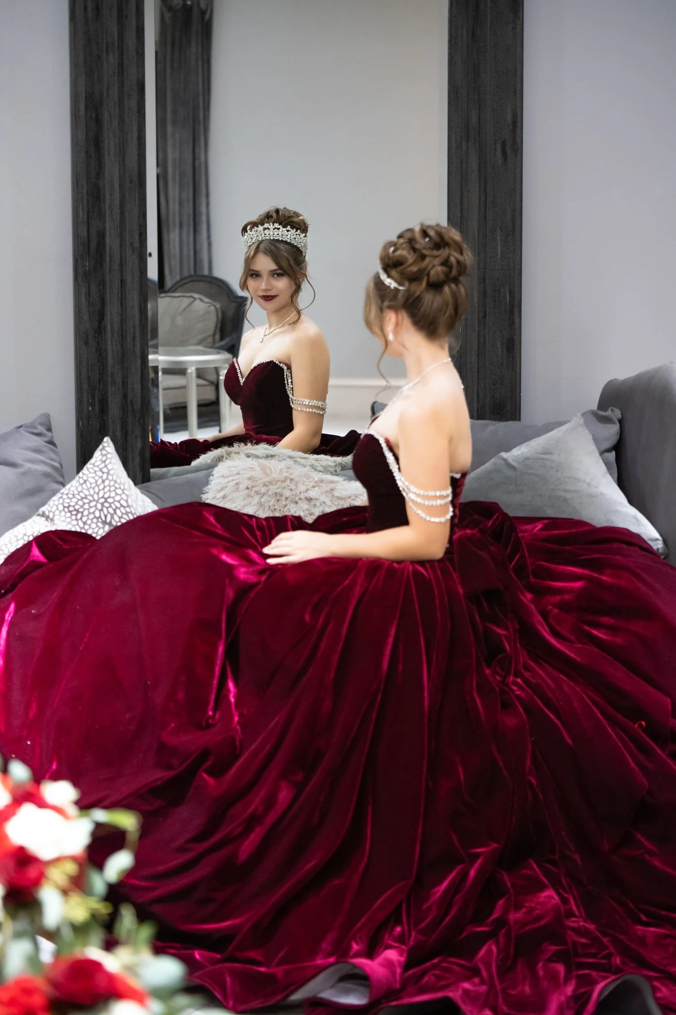 A breathtaking pre-reception portrait of Alana in her rich burgundy velvet Quinceañera gown. Seated gracefully with her reflection captured in the mirror, the image highlights her sparkling tiara and elegant styling. The dramatic flow of the gown cre