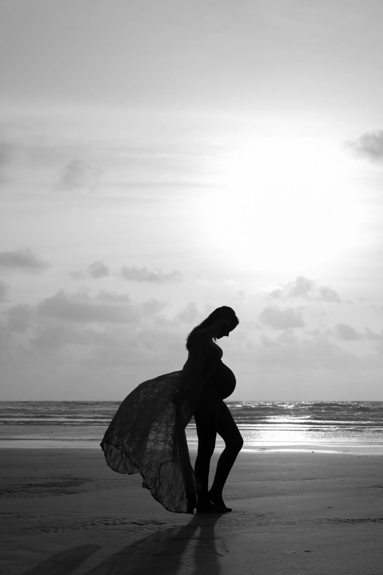 A black-and-white silhouette maternity portrait of an expecting mother walking barefoot along the shoreline at sunrise. The ocean reflects the soft light behind her, while a flowing sheer fabric trails in the wind, creating a dramatic and ethereal sc