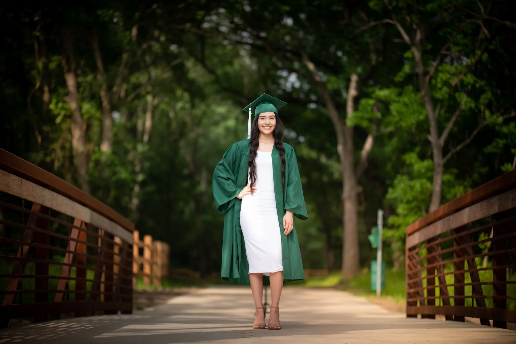 A full-length graduation portrait of a young woman wearing a green graduation gown over a white dress, standing confidently on a park bridge framed by tall trees.