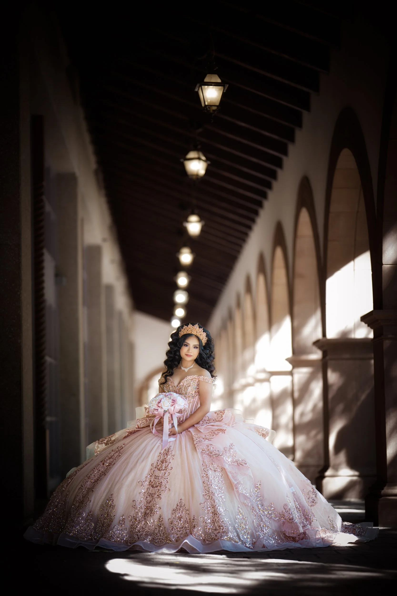 A quinceañera stands gracefully in a soft blush pink ball gown, holding a bouquet of pastel roses while surrounded by lush greenery. Natural light filters through the trees, creating a romantic and elegant portrait in an outdoor garden setting.