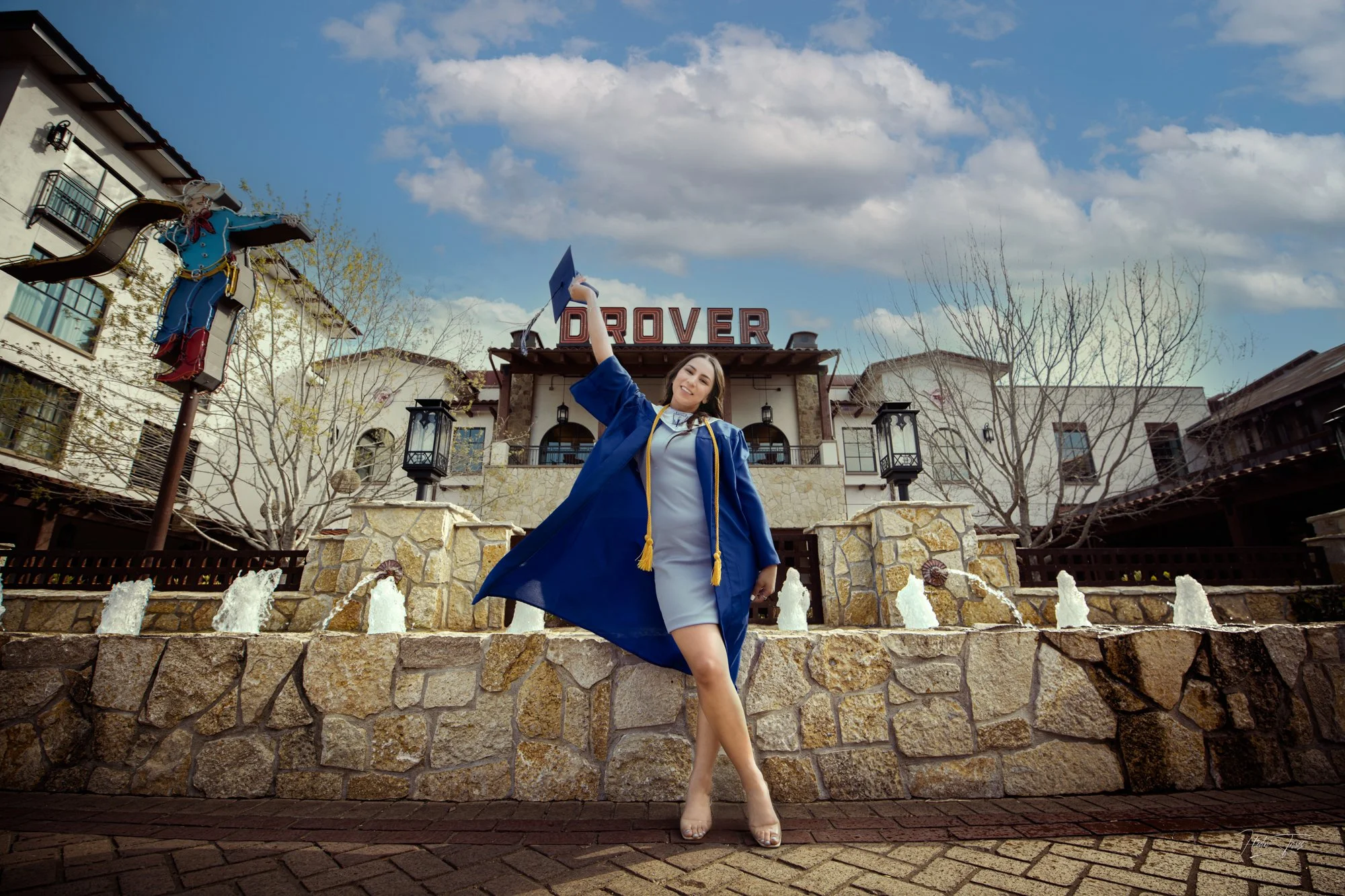 A graduate wearing a blue cap and gown celebrates in front of a stone fountain and rustic building, lifting her cap joyfully to mark the accomplishment.