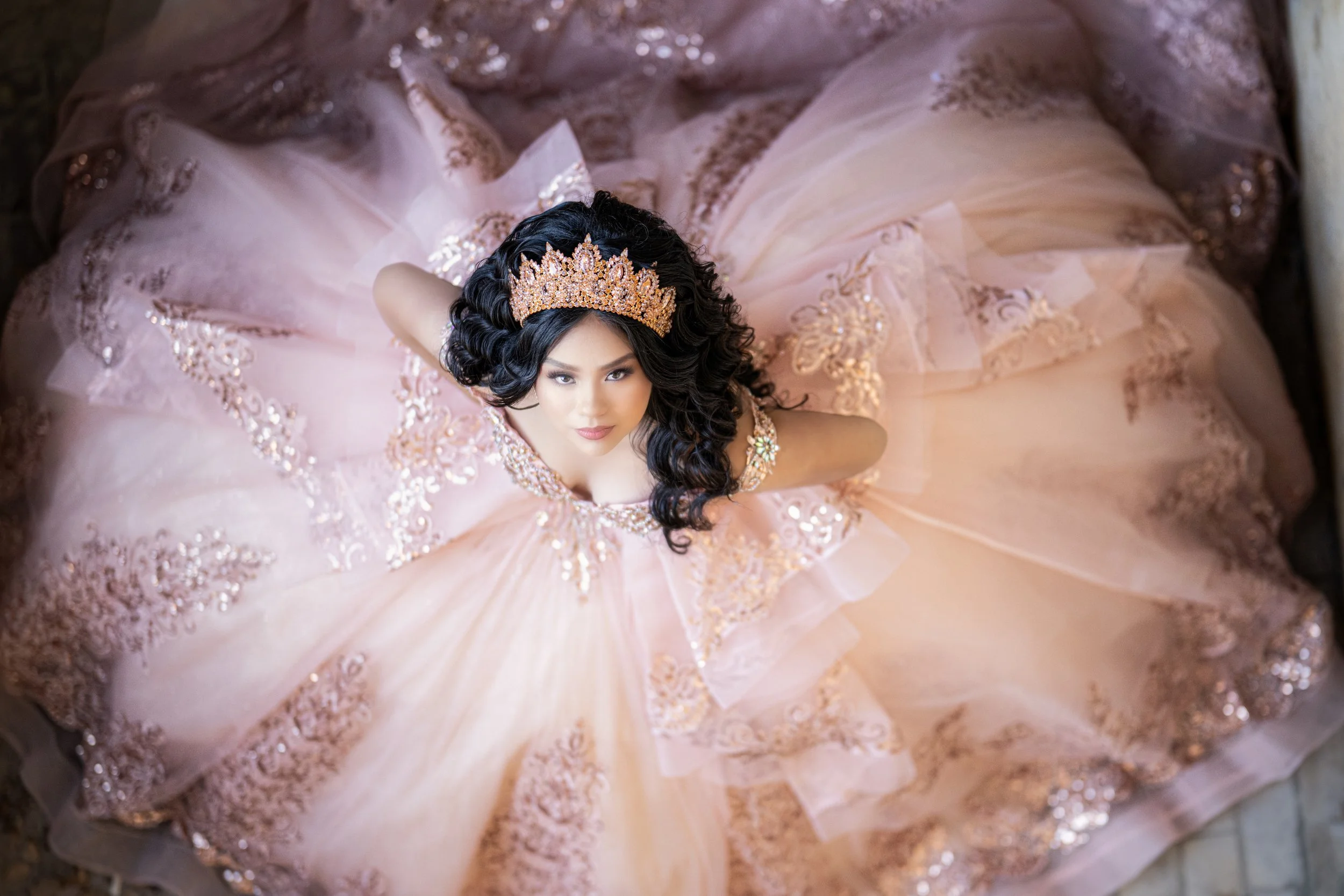 A dramatic overhead portrait of a quinceañera in a blush pink ball gown, captured from above as the dress fans out in a perfect circular shape. Intricate beadwork, embroidery, and layered tulle surround her as she gazes confidently into the camera, w