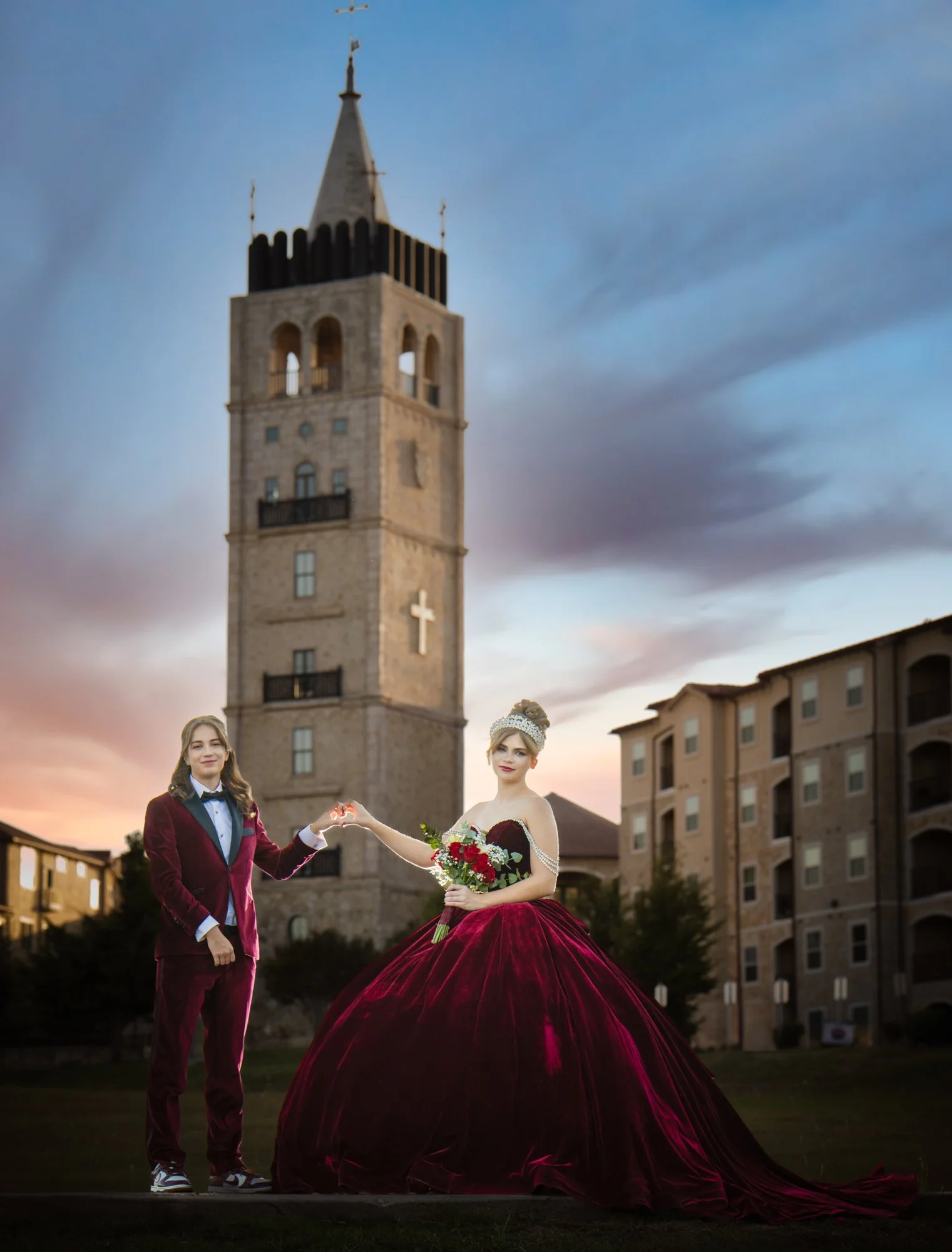 Twin quinceañeras stand hand in hand in front of a historic stone tower at sunset, wearing coordinated burgundy formal attire in a dramatic quinceañera portrait that blends tradition, elegance, and timeless architecture.