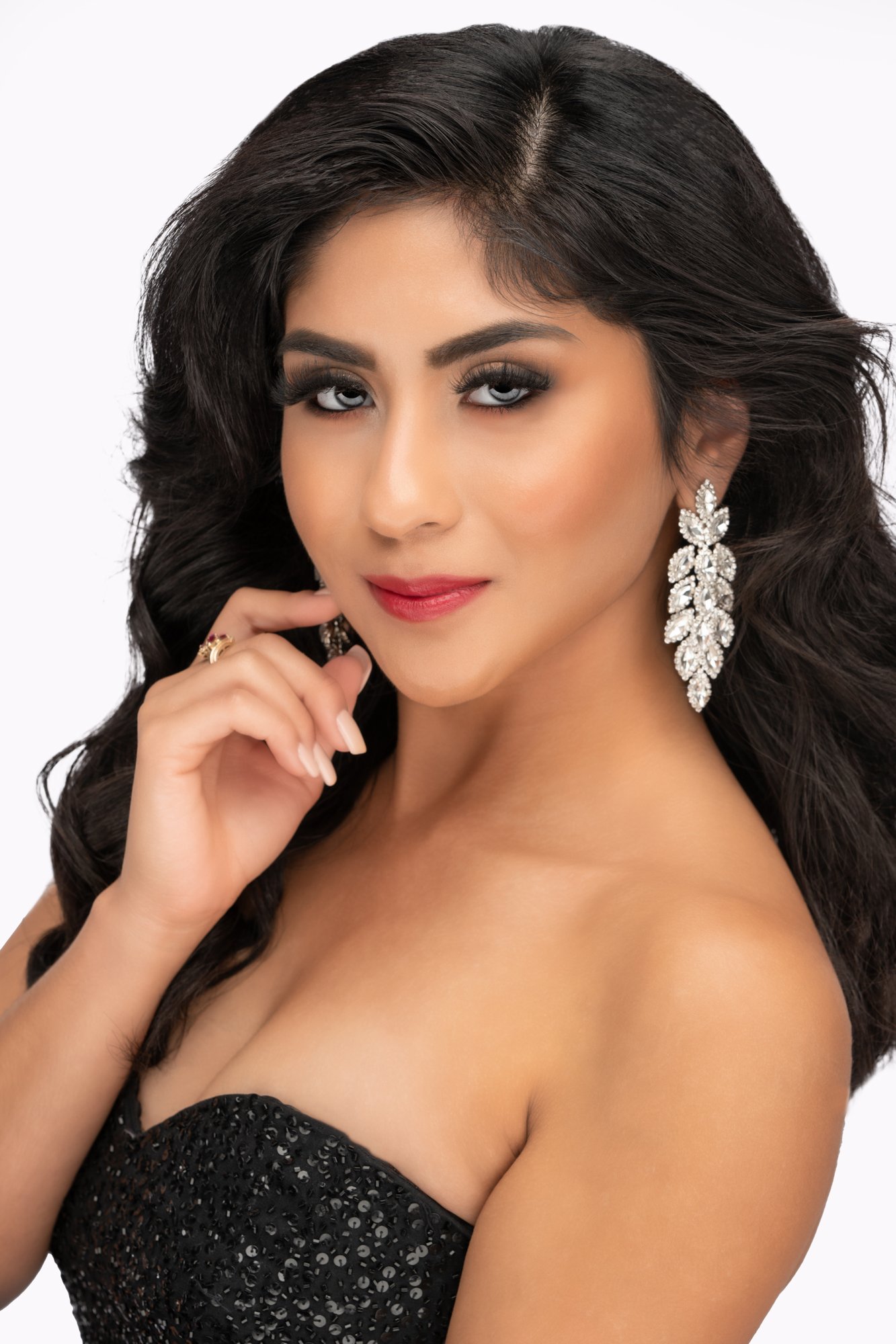A close-up studio beauty portrait of a young woman with long dark hair styled in soft waves, wearing dramatic crystal earrings and polished makeup, photographed against a white background with a confident, elegant expression.