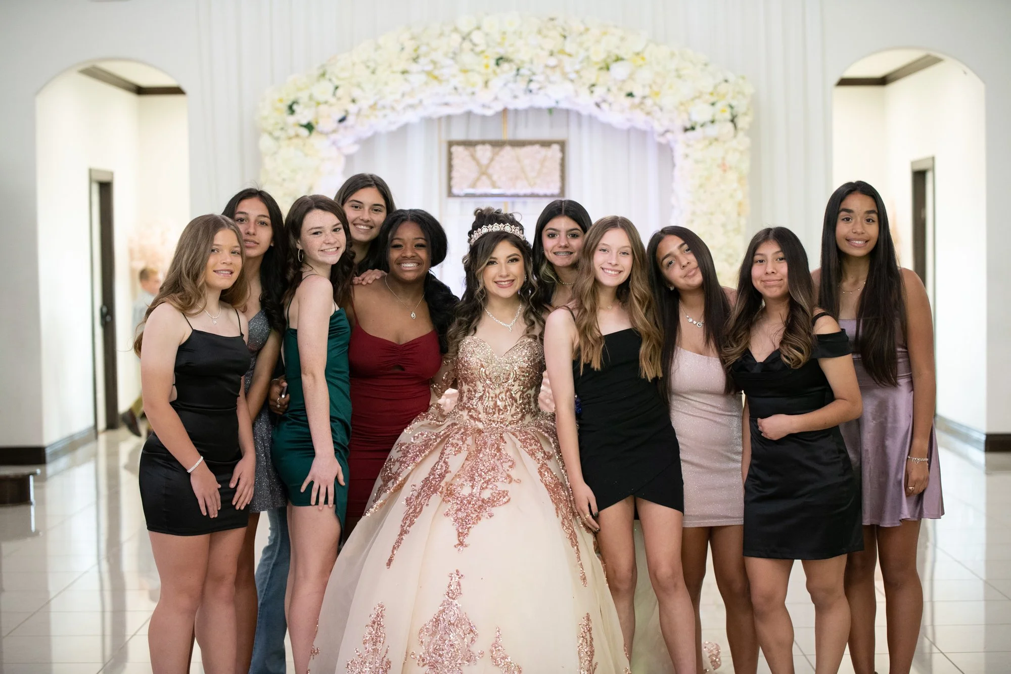Surrounded by her closest friends, Katelyn poses beneath a stunning floral archway. The neutral venue palette and soft backdrop highlight the warmth, laughter, and bond shared between lifelong friends.