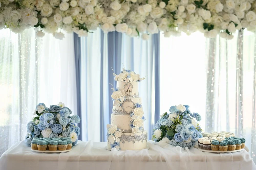 Luxury blue and white Quinceañera cake display featuring a four-tier custom cake, floral arrangements, cupcakes, and elegant reception décor under a floral canopy.