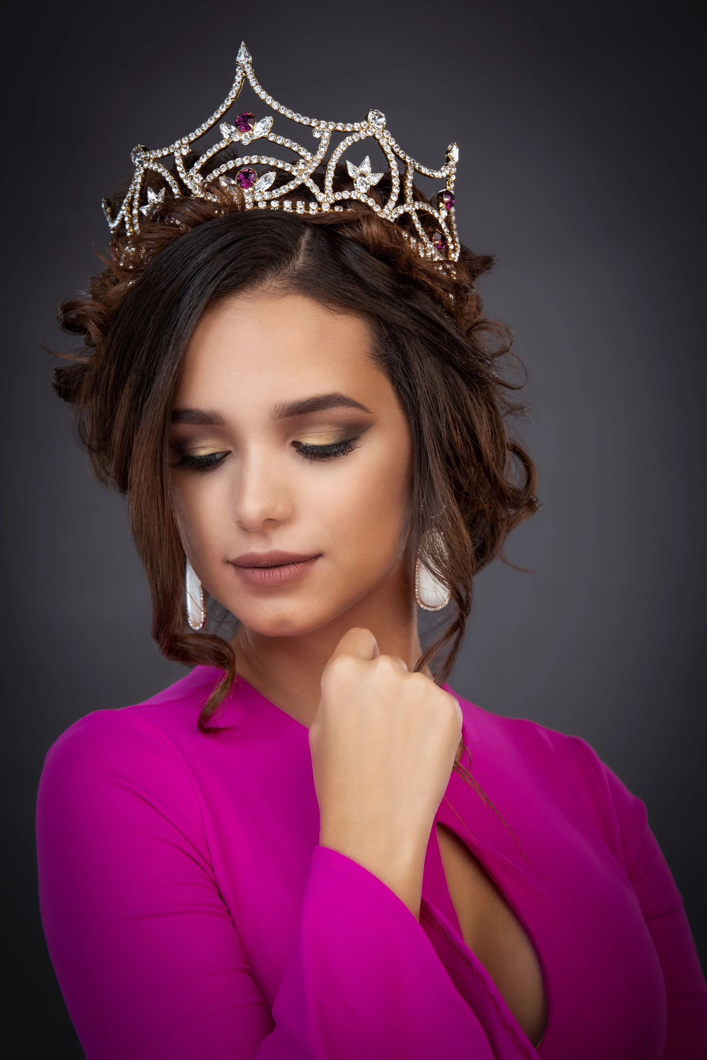 A close-up studio portrait of the same crowned woman in a magenta dress, captured in a soft, elegant pose with her gaze lowered and her hand gently resting near her chin. The jeweled crown and refined makeup are highlighted, creating a timeless, rega
