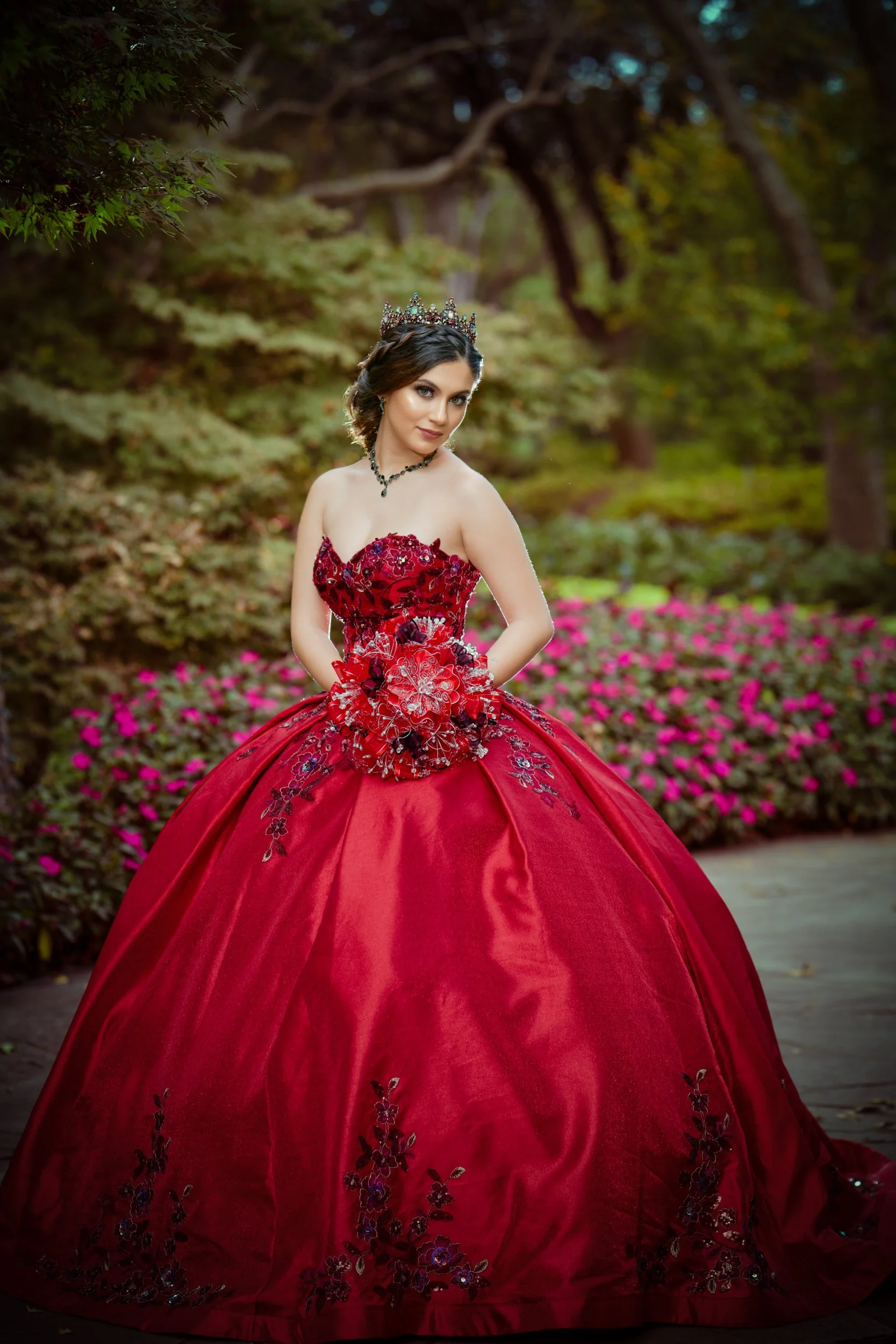 Quinceañera portrait of a young woman in a red gown and crown outdoors.
