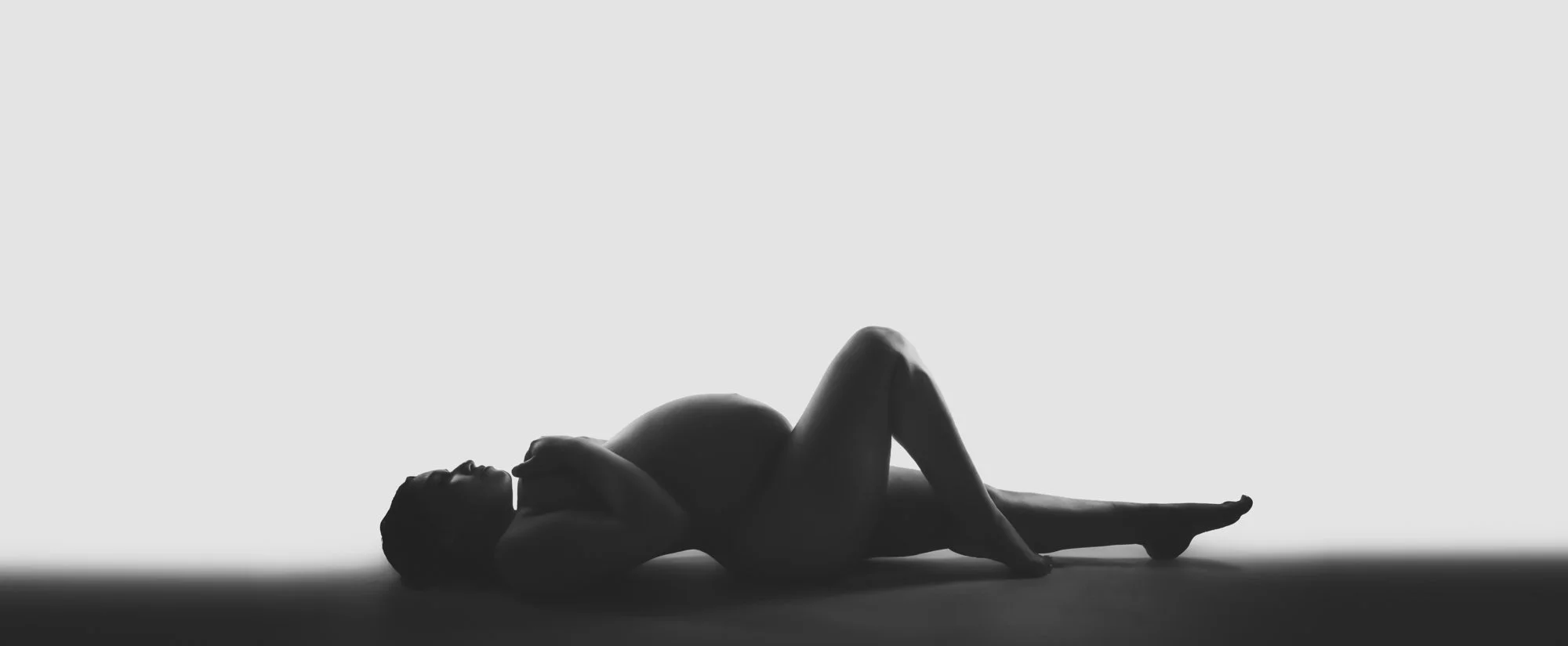 A minimalist black-and-white maternity fine-art portrait of an expecting mother reclining gracefully. The dramatic side lighting accentuates the natural curves of pregnancy against a clean white background, creating a sculptural and timeless composit