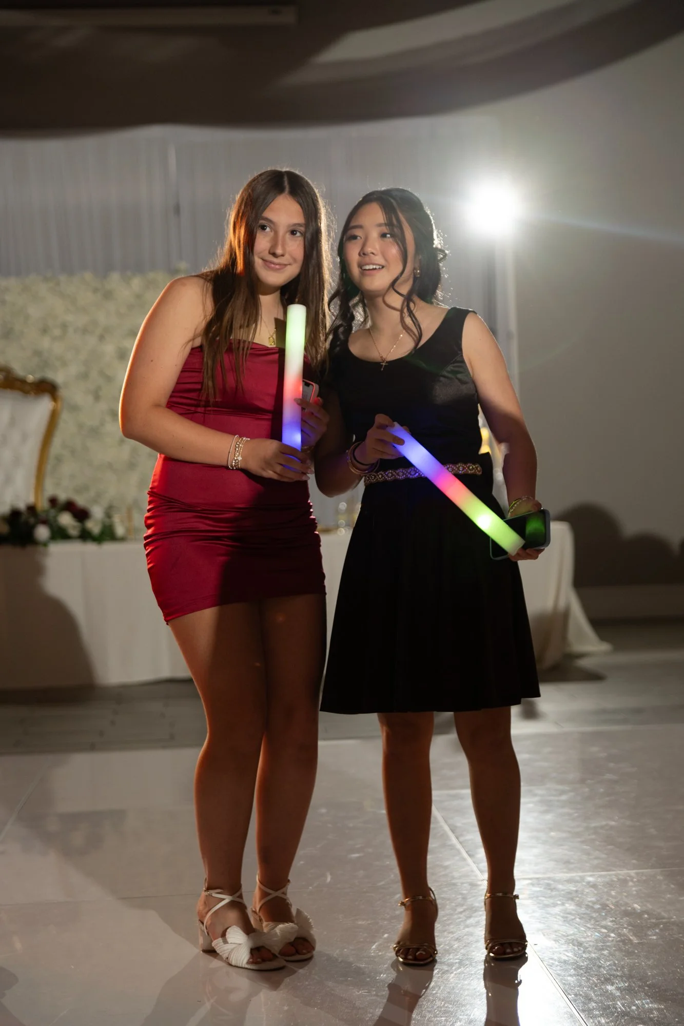 Two guests pause for a candid moment holding colorful glow sticks as the party lights illuminate the reception space. The image captures the playful, youthful energy that keeps the celebration alive all night long.