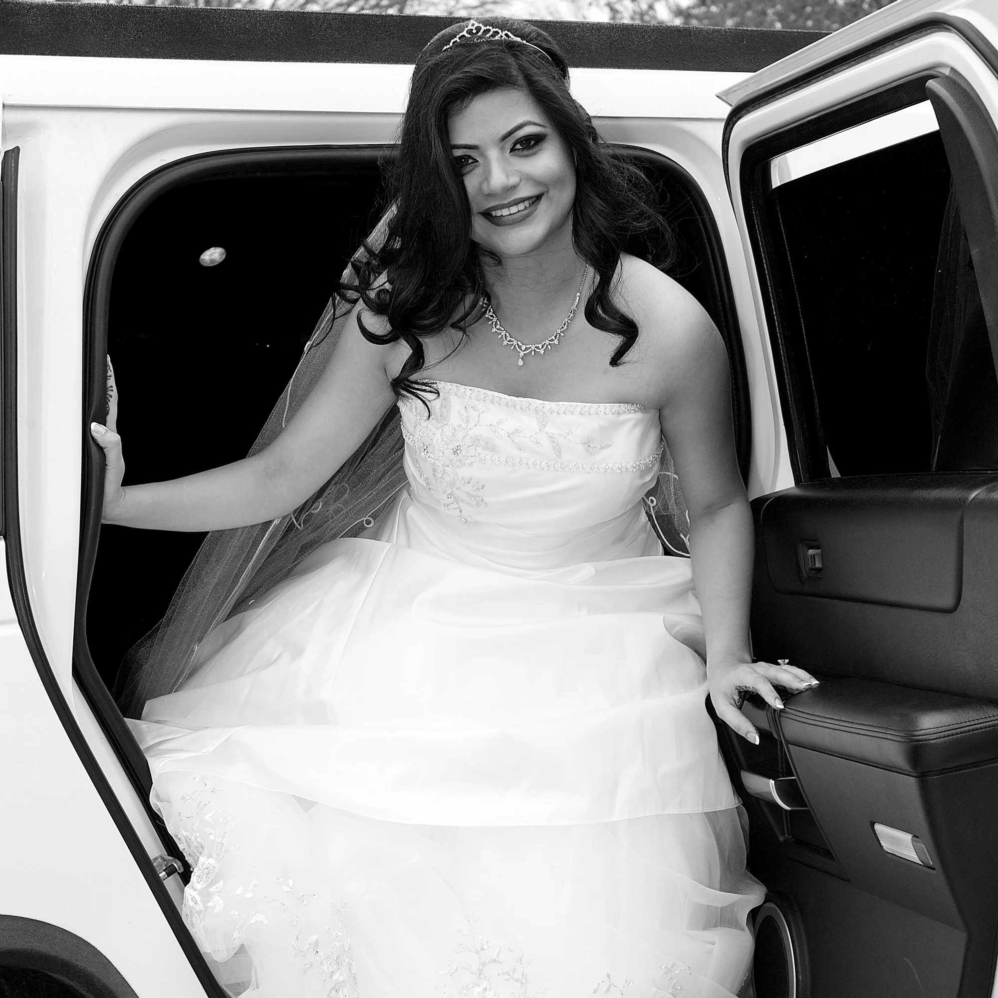 Smiling bride stepping gracefully out of a luxury vehicle, dressed in a strapless wedding gown with a flowing veil, captured in a timeless black and white bridal portrait.
