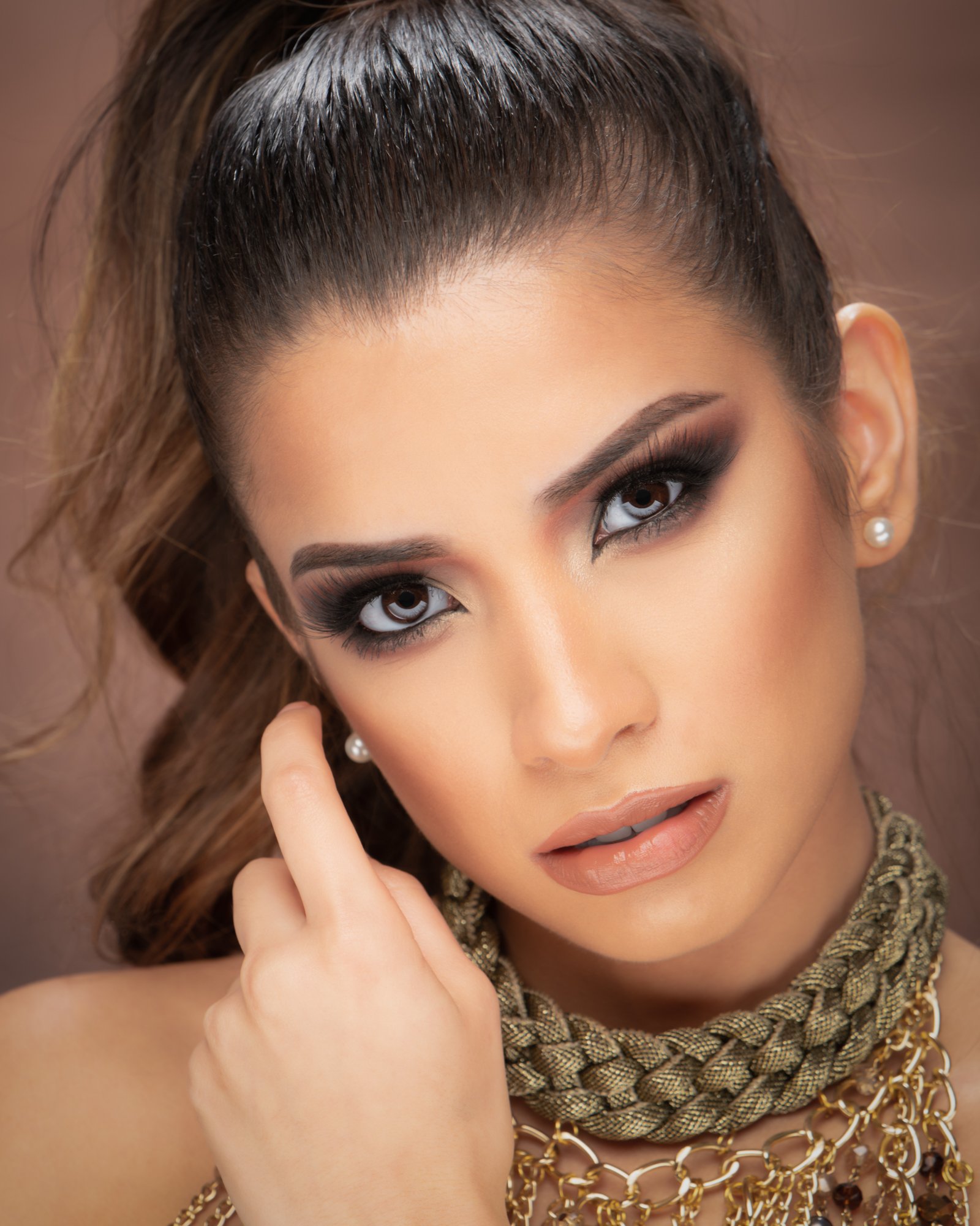 A polished studio portrait featuring glamorous makeup, soft lighting, and a warm neutral backdrop. The subject gazes directly at the camera, wearing layered statement jewelry and pearl earrings for a refined look.