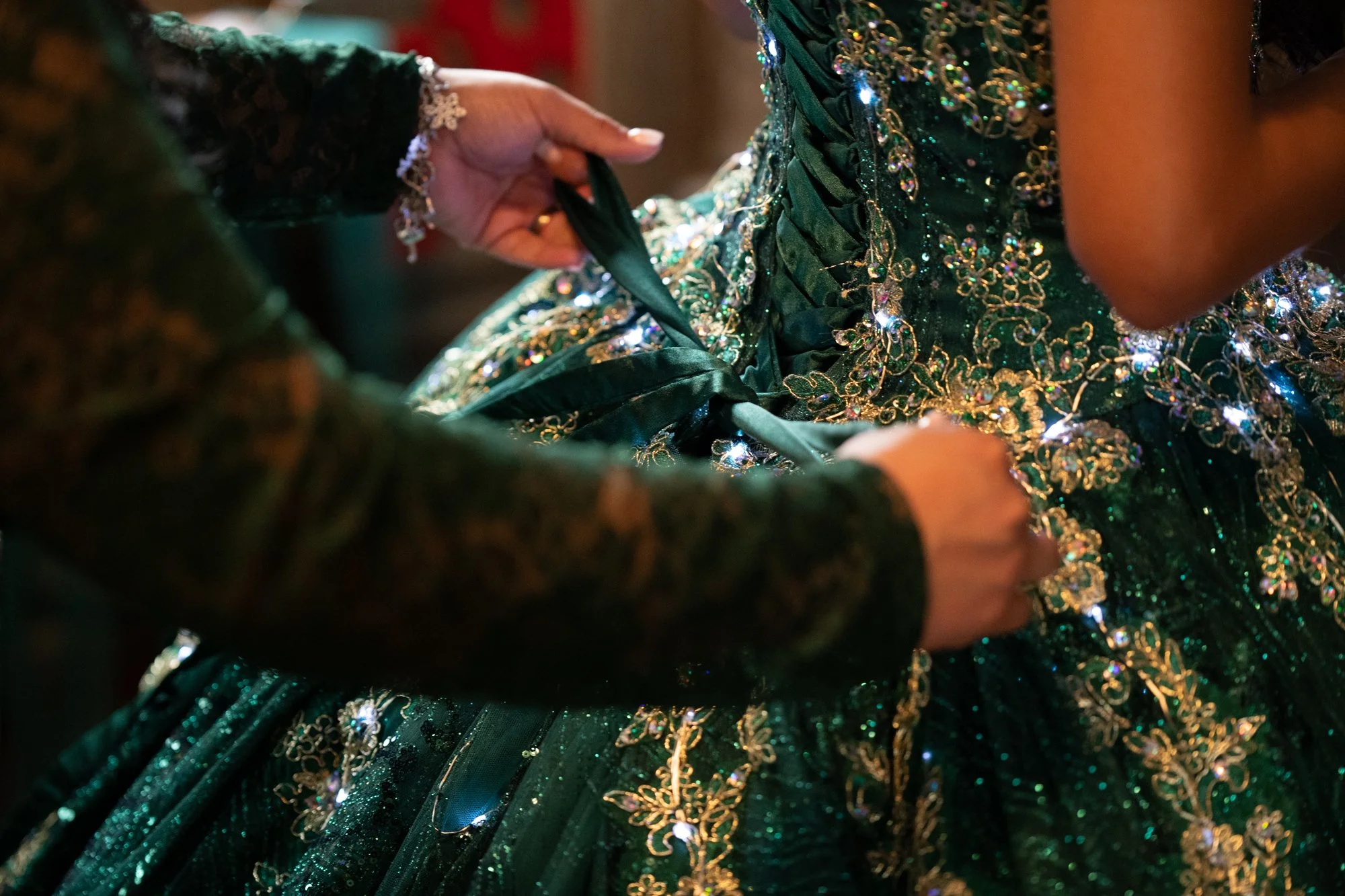 A close-up moment as the gown is carefully tightened and secured. The rich emerald fabric, gold embroidery, and shimmering embellishments catch the light beautifully, symbolizing the final transformation from daughter to Quinceañera.