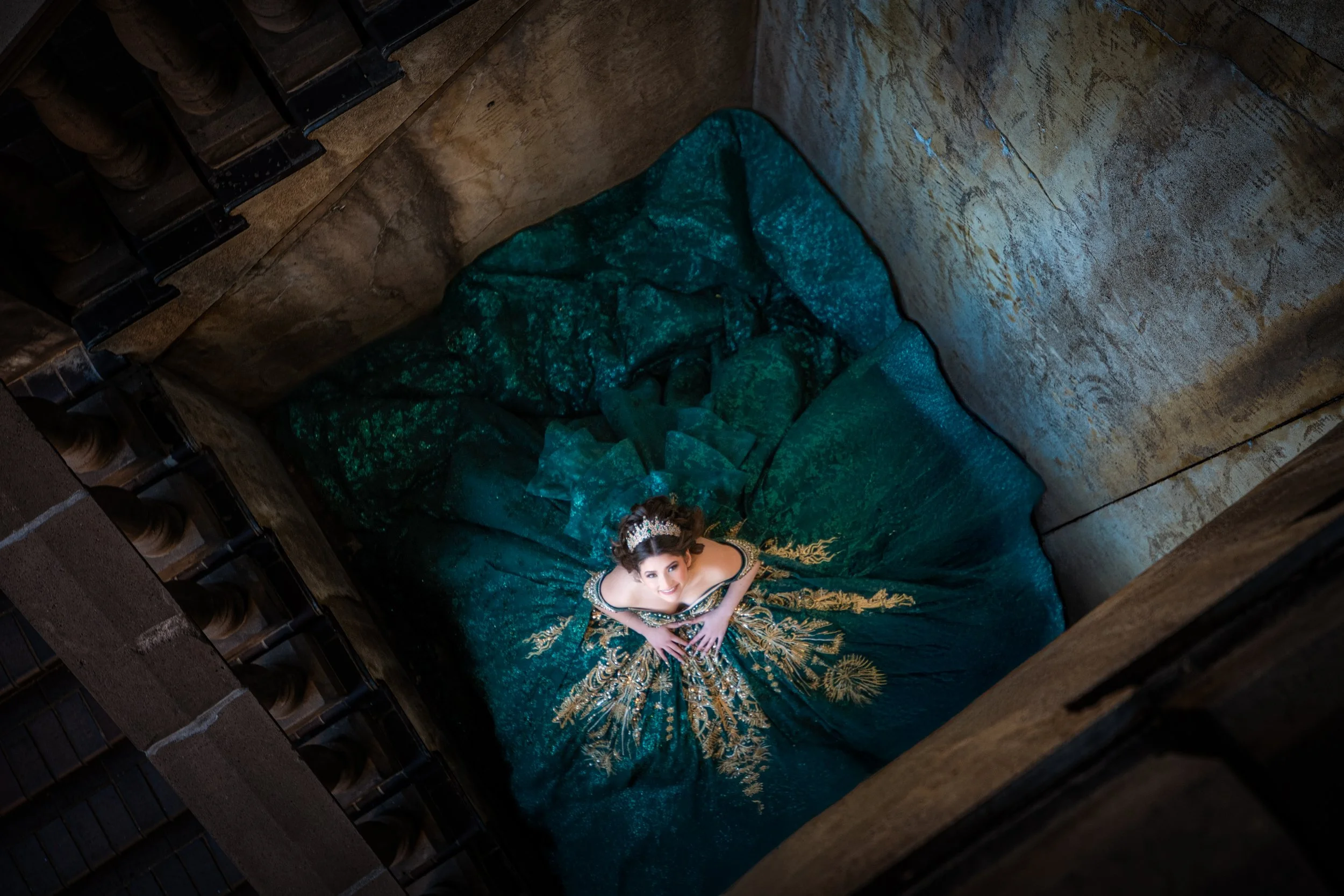 A top-down artistic Quinceañera portrait capturing the full dramatic spread of an emerald green ball gown arranged in a circular pattern, emphasizing texture, sparkle, and symmetry.