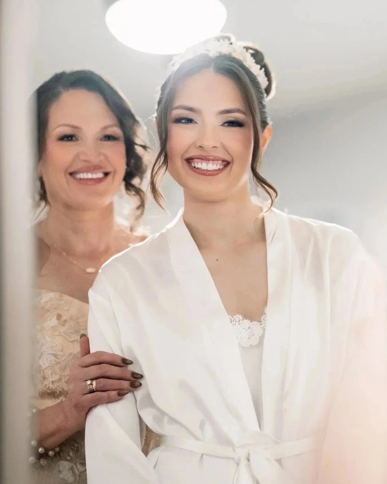 A bride smiling in a white robe during bridal prep, standing in front of a mirror with her mother behind her, sharing a joyful and emotional moment before the wedding ceremony.