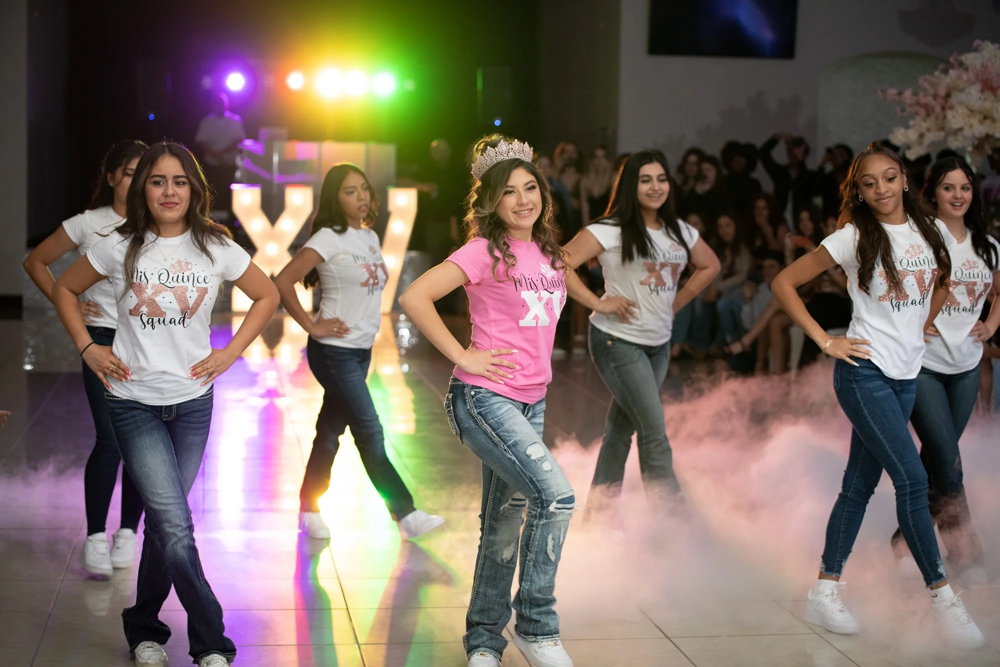Under colorful stage lighting and low-lying fog, Katelyn and her squad strike a synchronized pose in matching custom tees and denim. The illuminated “XV” letters glow in the background, adding a bold statement to this modern Quinceañera celebration.