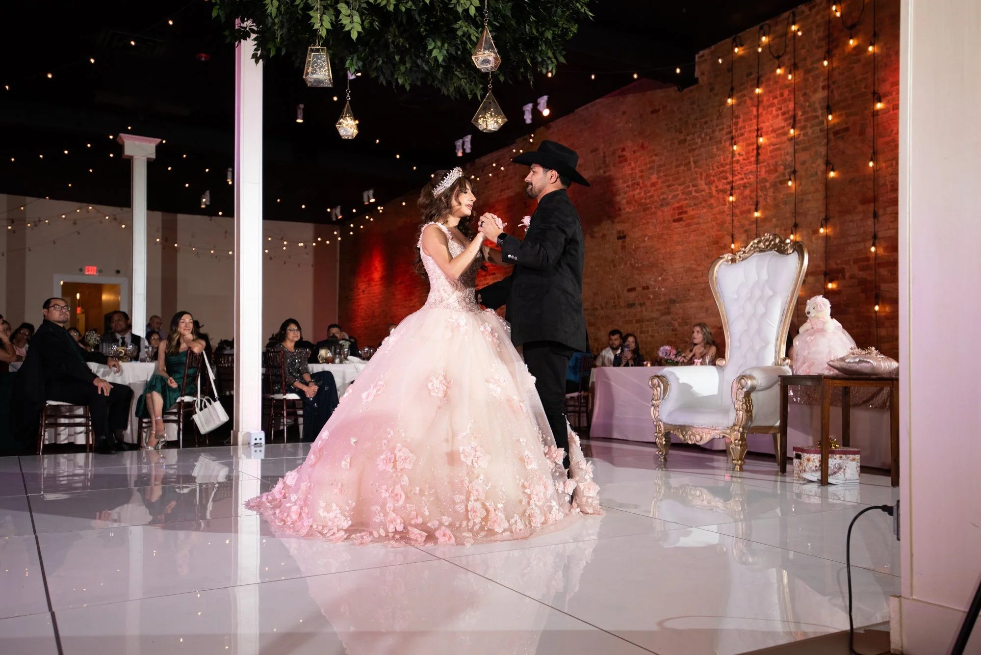 Under twinkling lights and warm ambient glow, she shares a graceful dance with her father. The sweeping blush gown reflects beautifully across the glossy dance floor, capturing both elegance and emotion.
