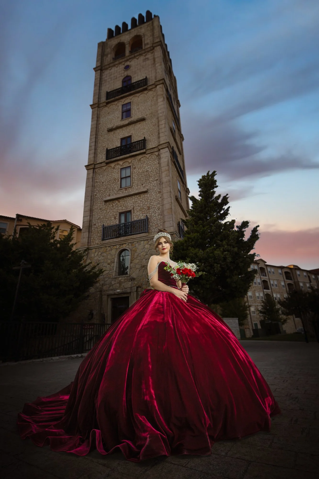 A quinceañera in a luxurious burgundy ball gown poses confidently beneath a towering stone bell tower at dusk, creating a dramatic and elegant quinceañera portrait that highlights grace, beauty, and architectural grandeur.