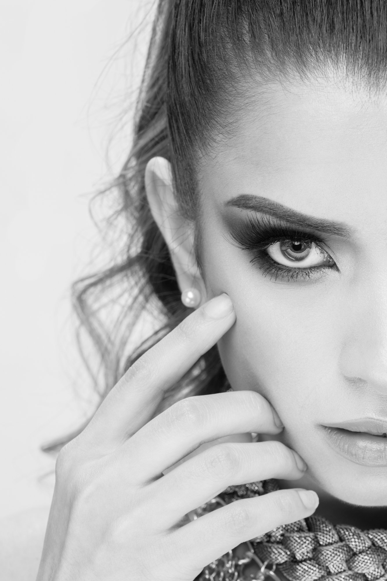 A high-contrast black-and-white portrait highlighting one side of the subject’s face. Dramatic eye makeup, long lashes, and elegant hand placement draw attention to the eye and facial structure.