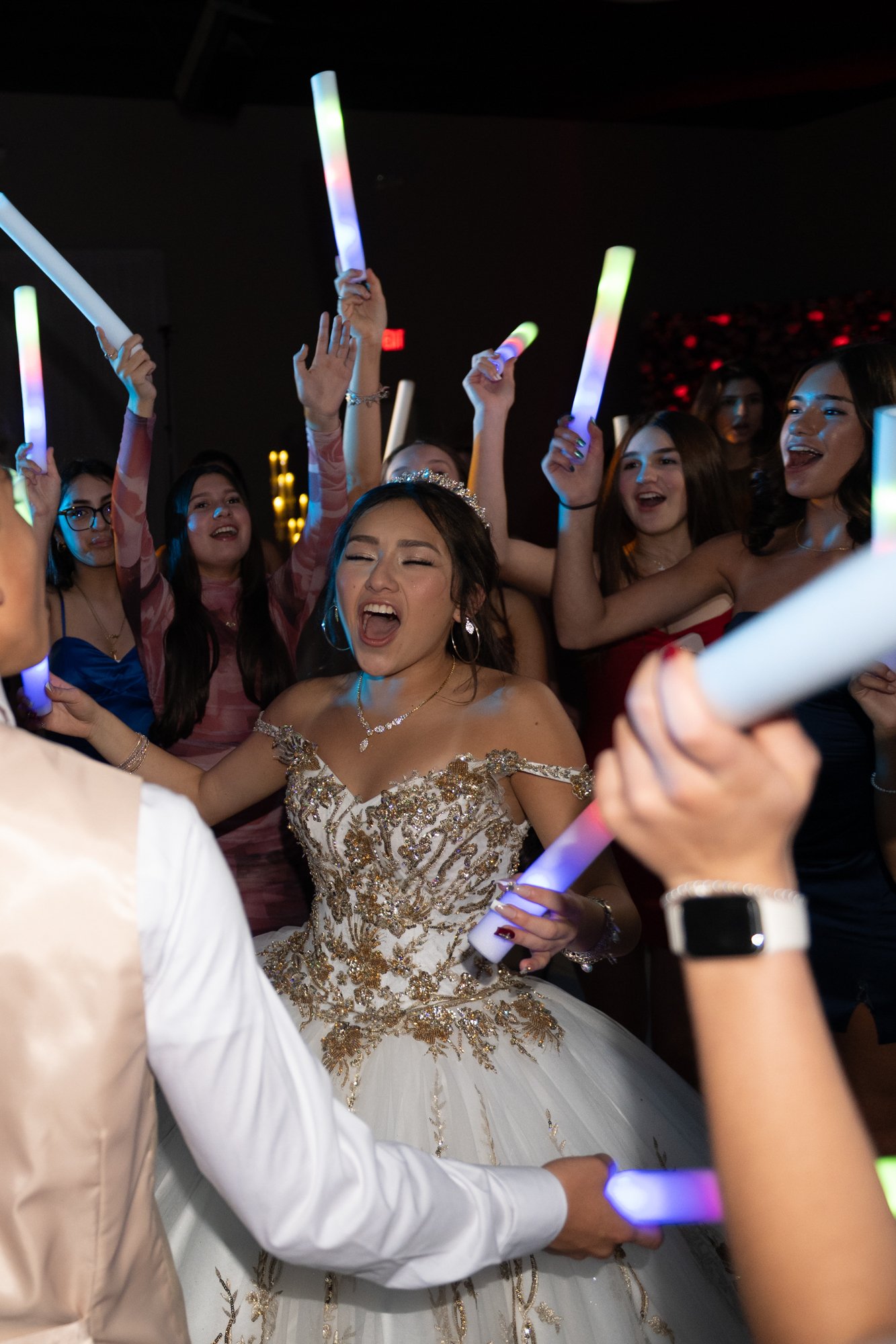 The energy peaks as Alex celebrates surrounded by friends waving glowing party sticks in the air. Smiles, laughter, and vibrant lights capture the pure excitement and joyful chaos that define an unforgettable Quinceañera dance floor moment.