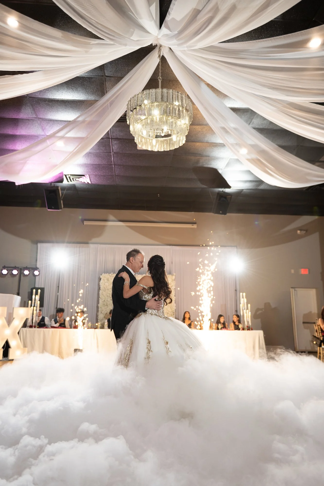A breathtaking dance floor scene as Alex shares a special father-daughter dance surrounded by low-lying fog and sparkling cold sparklers. The chandelier overhead and glowing lights create a magical atmosphere, turning this tradition into a cinematic,
