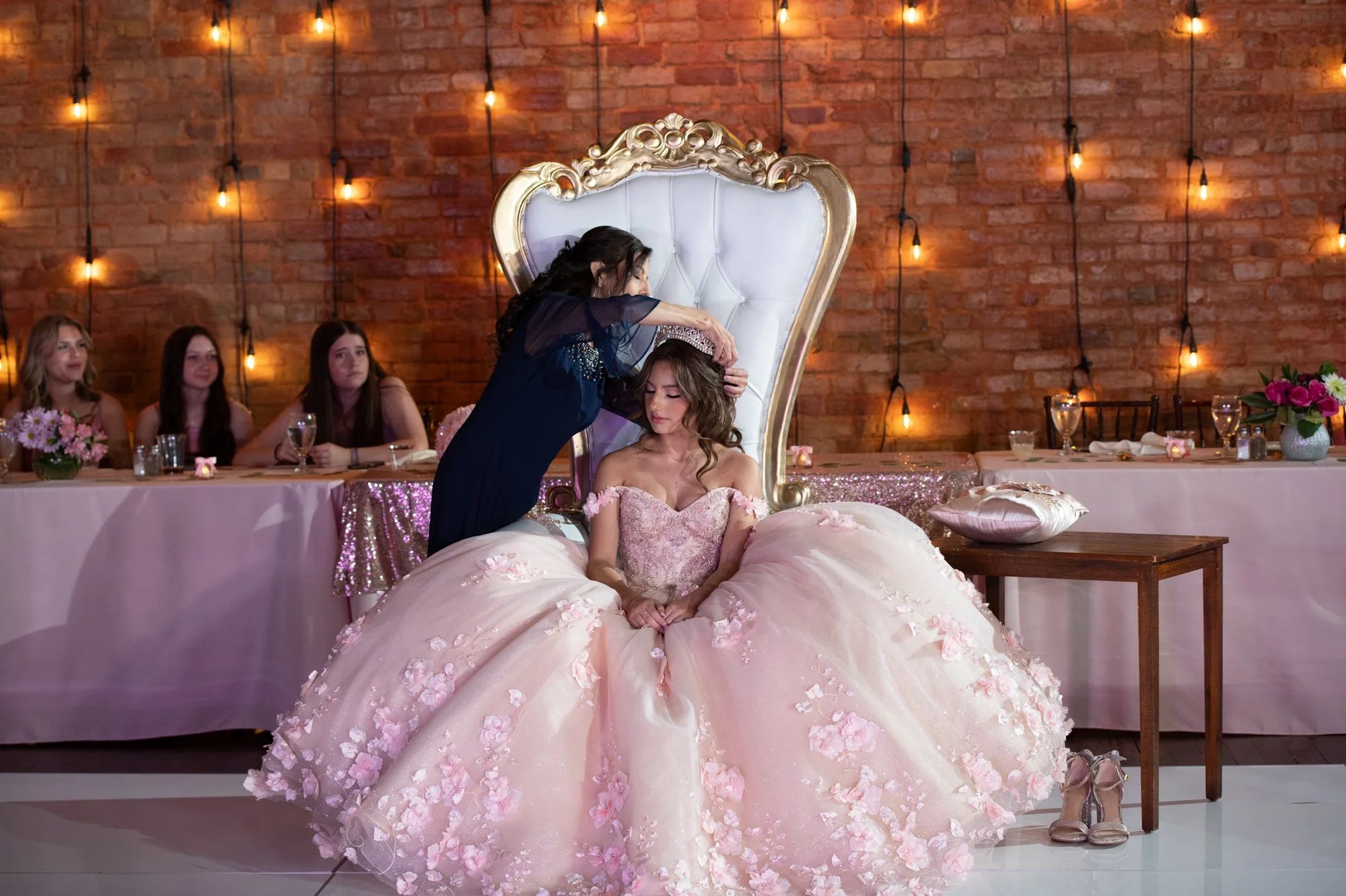Seated on an elegant throne against warm brick and string lights, the quinceañera bows her head as someone carefully places her crown. A powerful ceremonial moment filled with symbolism and tradition.