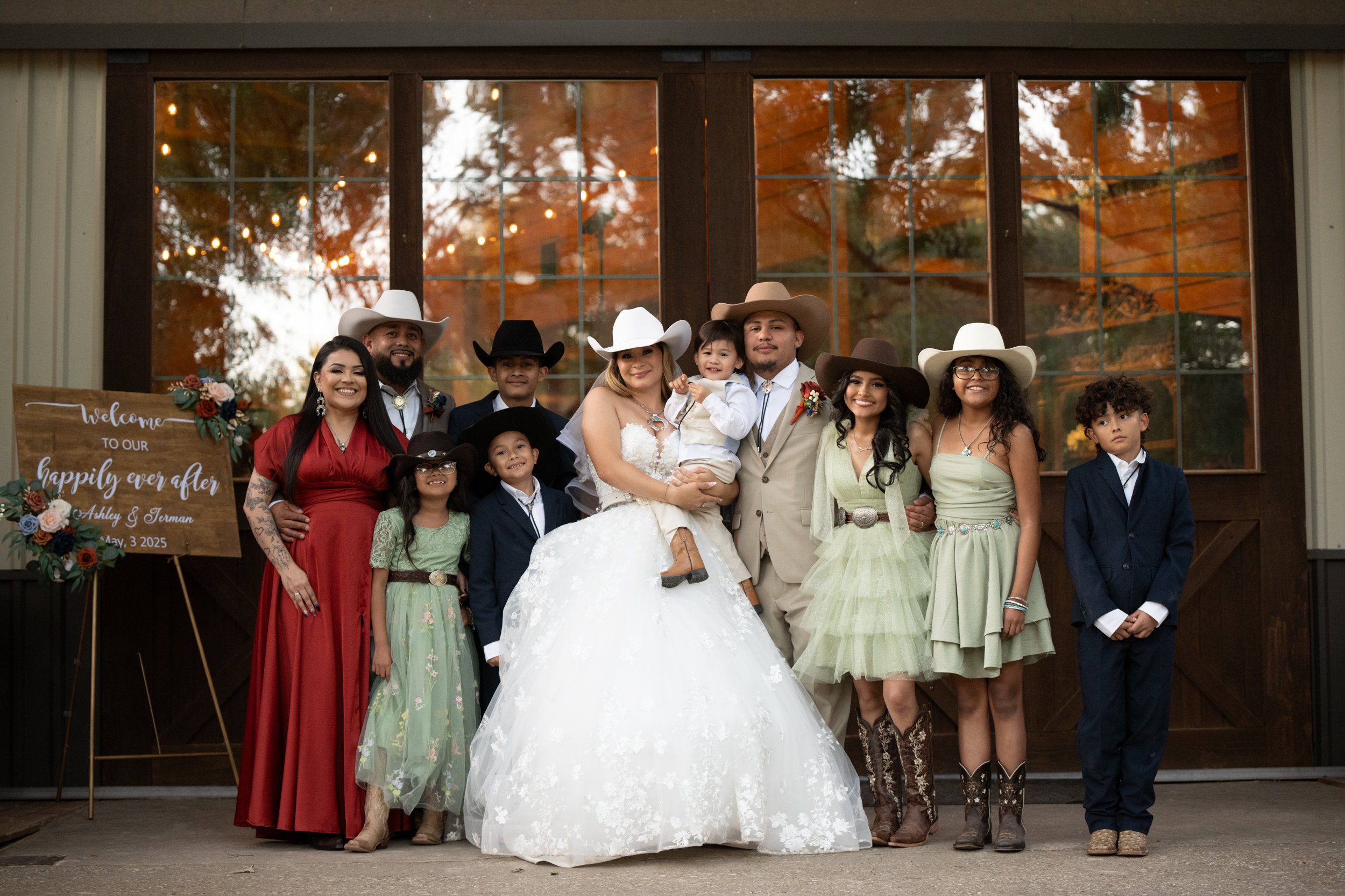 The bride and groom stand proudly surrounded by their wedding party and family, blending rustic elegance and heartfelt connection in a joyful group portrait filled with warmth and celebration.