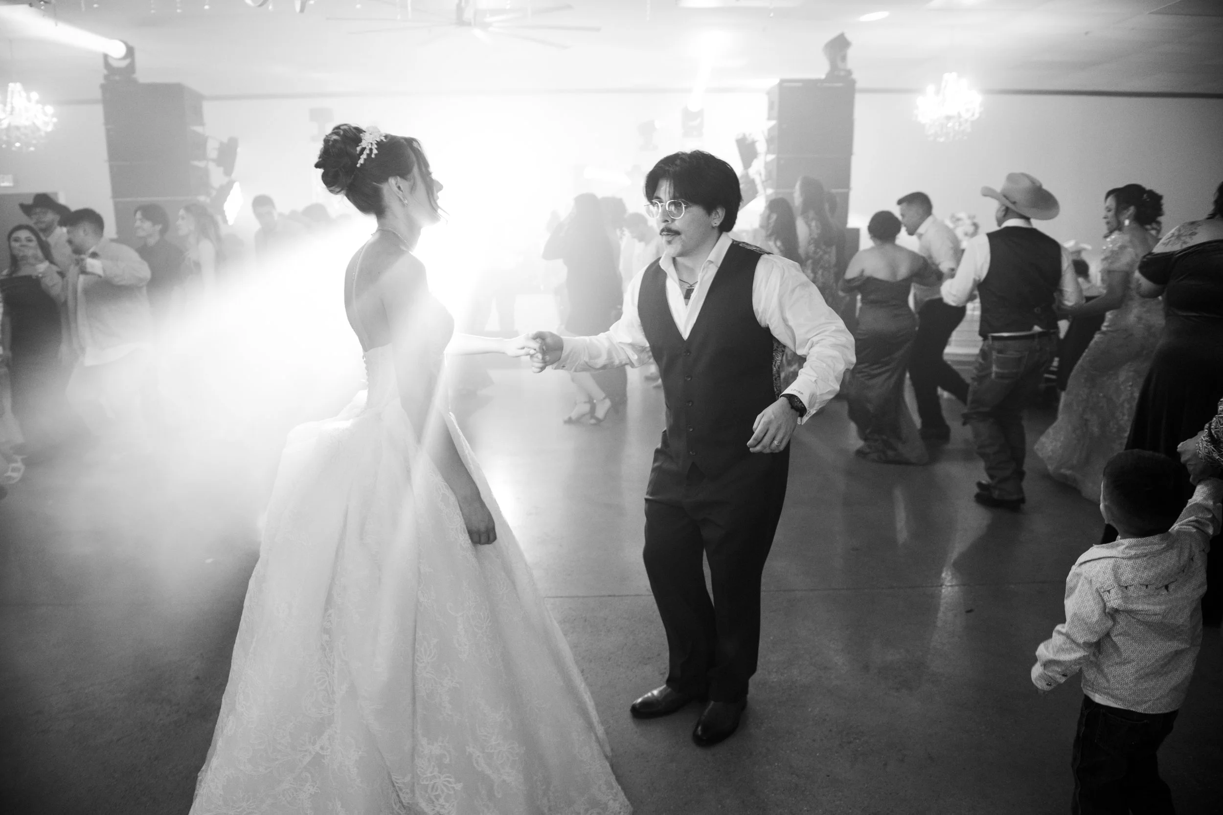 A dramatic black-and-white reception photo of the bride dancing under beams of light on a crowded dance floor, creating a cinematic and energetic atmosphere.