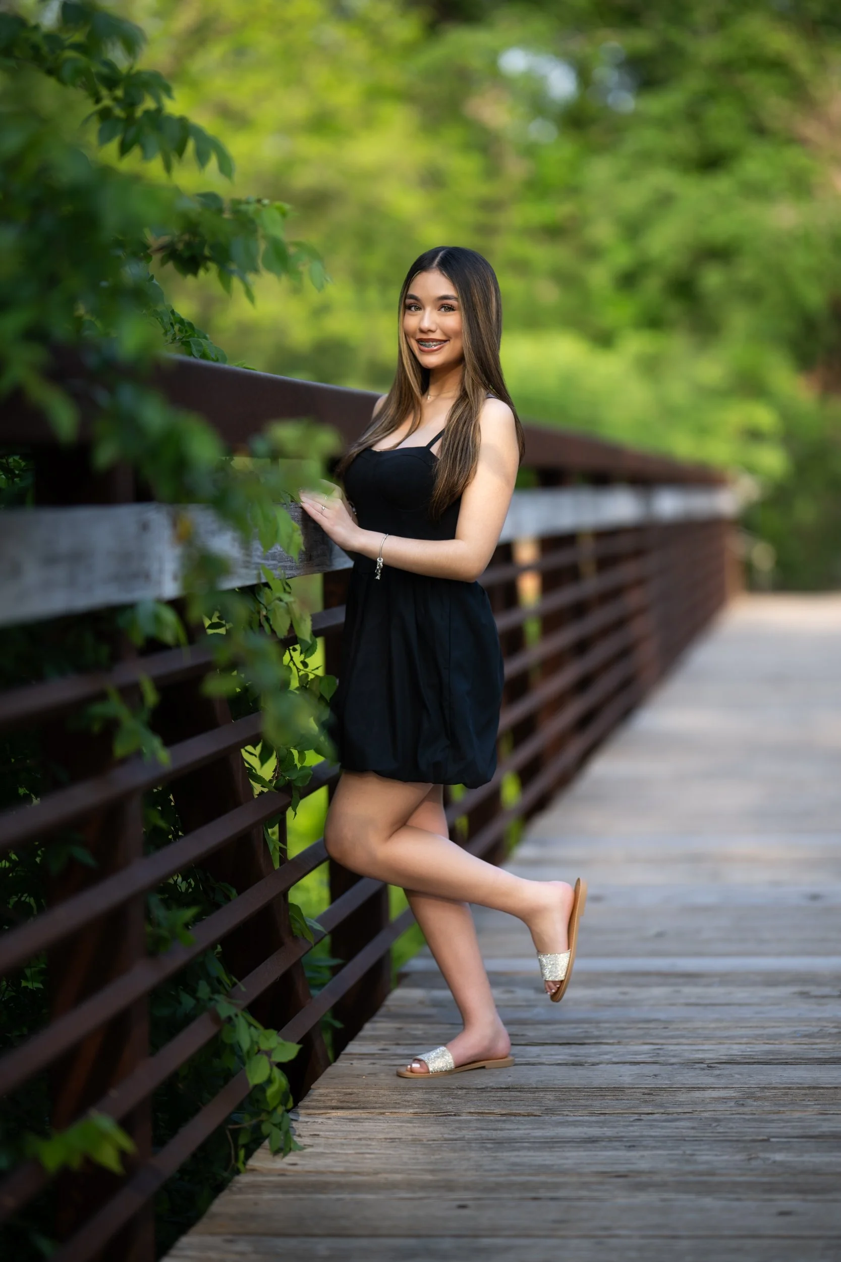 A young woman wearing a black sleeveless dress poses on a wooden pedestrian bridge surrounded by lush green trees, smiling softly as natural light highlights the background.