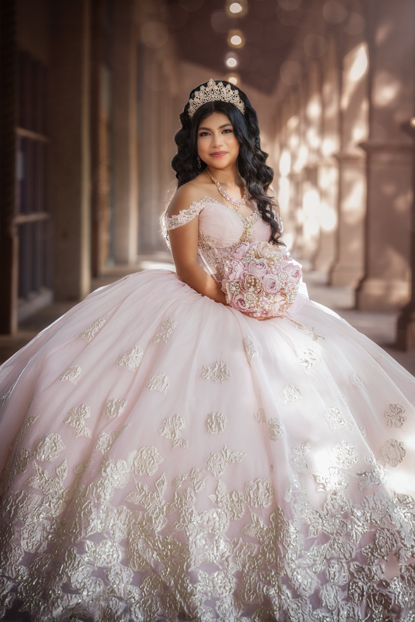 The quinceañera stands gracefully beneath an arched stone hallway, wearing a blush pink ball gown adorned with delicate embroidery. Holding a floral bouquet close to her waist, she gazes confidently toward the camera as soft, warm light filters throu