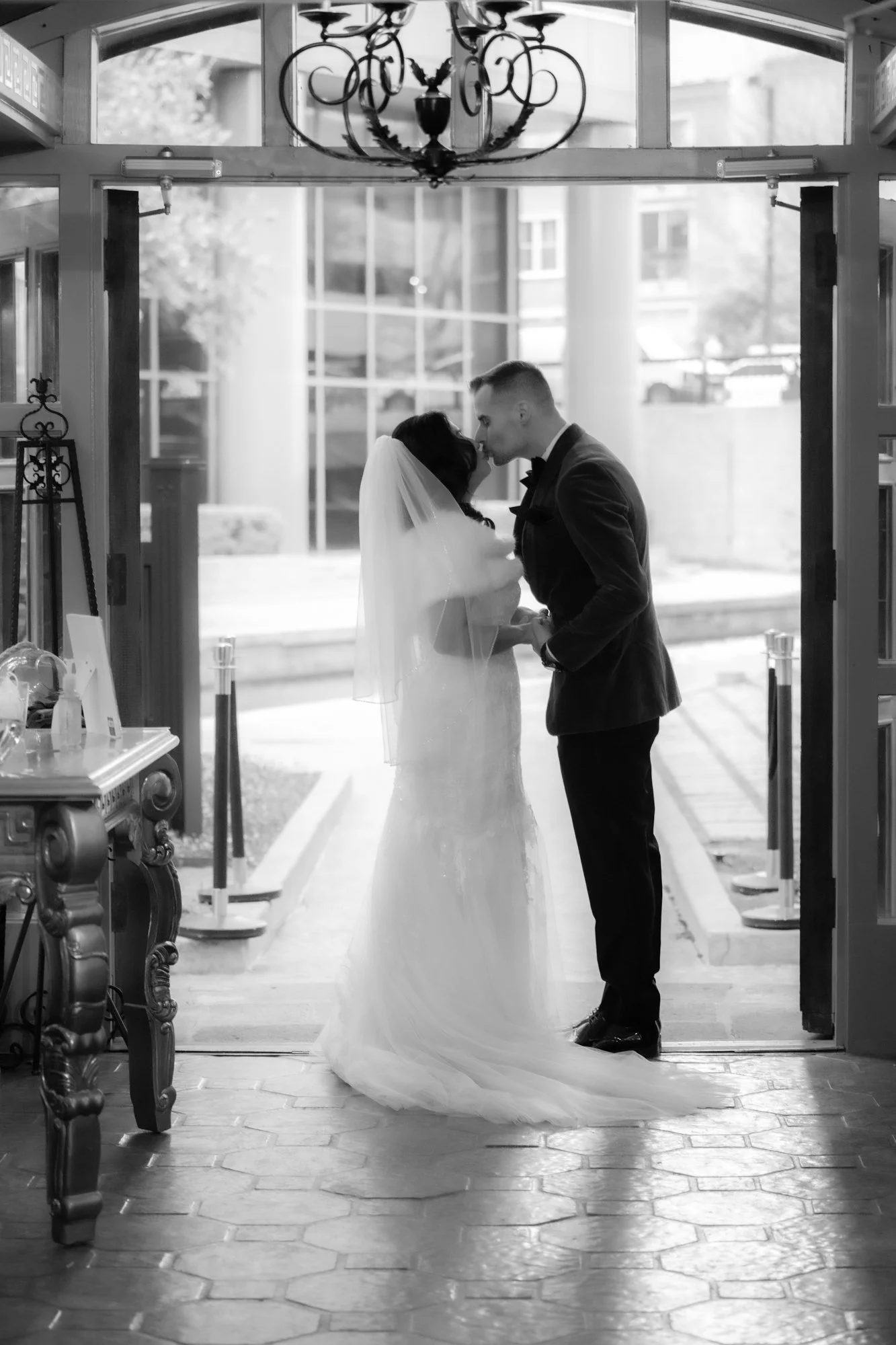 Black and white wedding portrait of a bride and groom sharing a kiss beneath an elegant chandelier at the venue entrance, framed by open doors and soft natural light.
