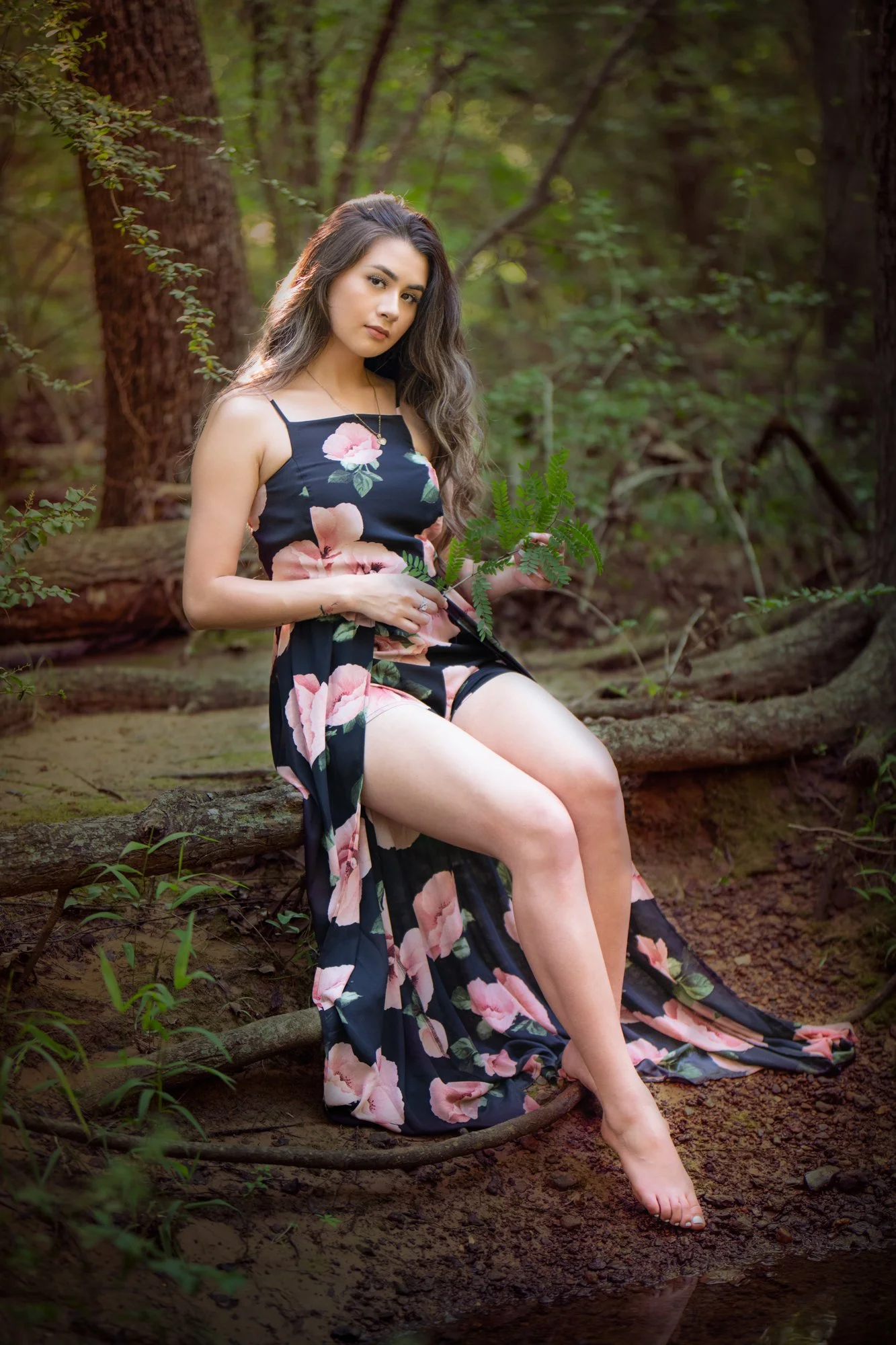A relaxed portrait of a woman sitting on fallen logs in a forest setting, holding greenery in her hands. She wears a black floral dress and looks calmly toward the camera, surrounded by soft natural light and earthy textures.