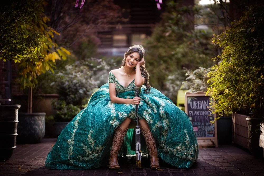 Natalie Vega smiles while seated in a charming urban garden setting, wearing a stunning emerald green Quinceañera gown and cowboy boots. This portrait perfectly blends Texas charm with timeless Quinceañera elegance.