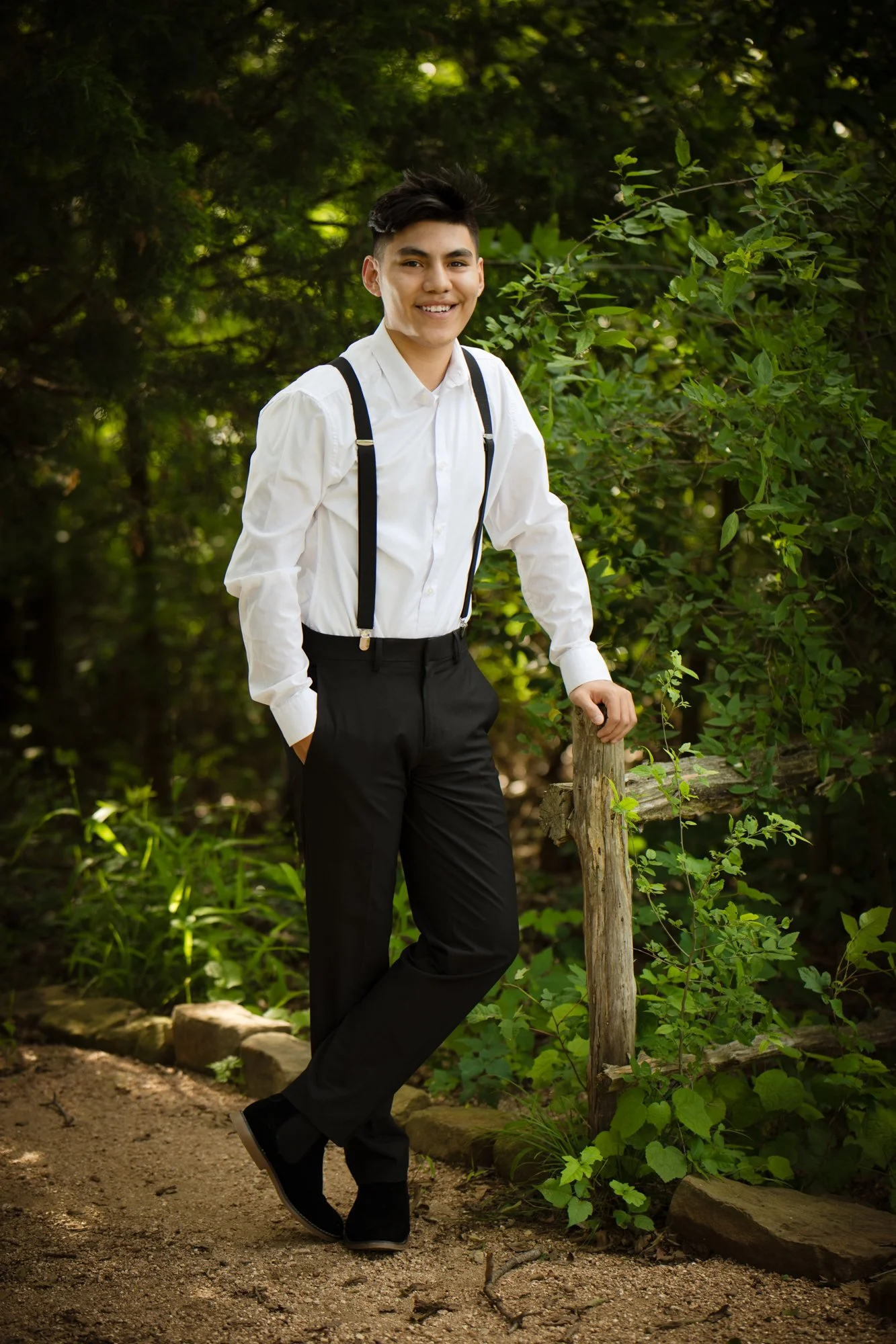 A young man wearing a white shirt with black suspenders stands in a wooded outdoor setting, smiling confidently while leaning against a wooden post.
