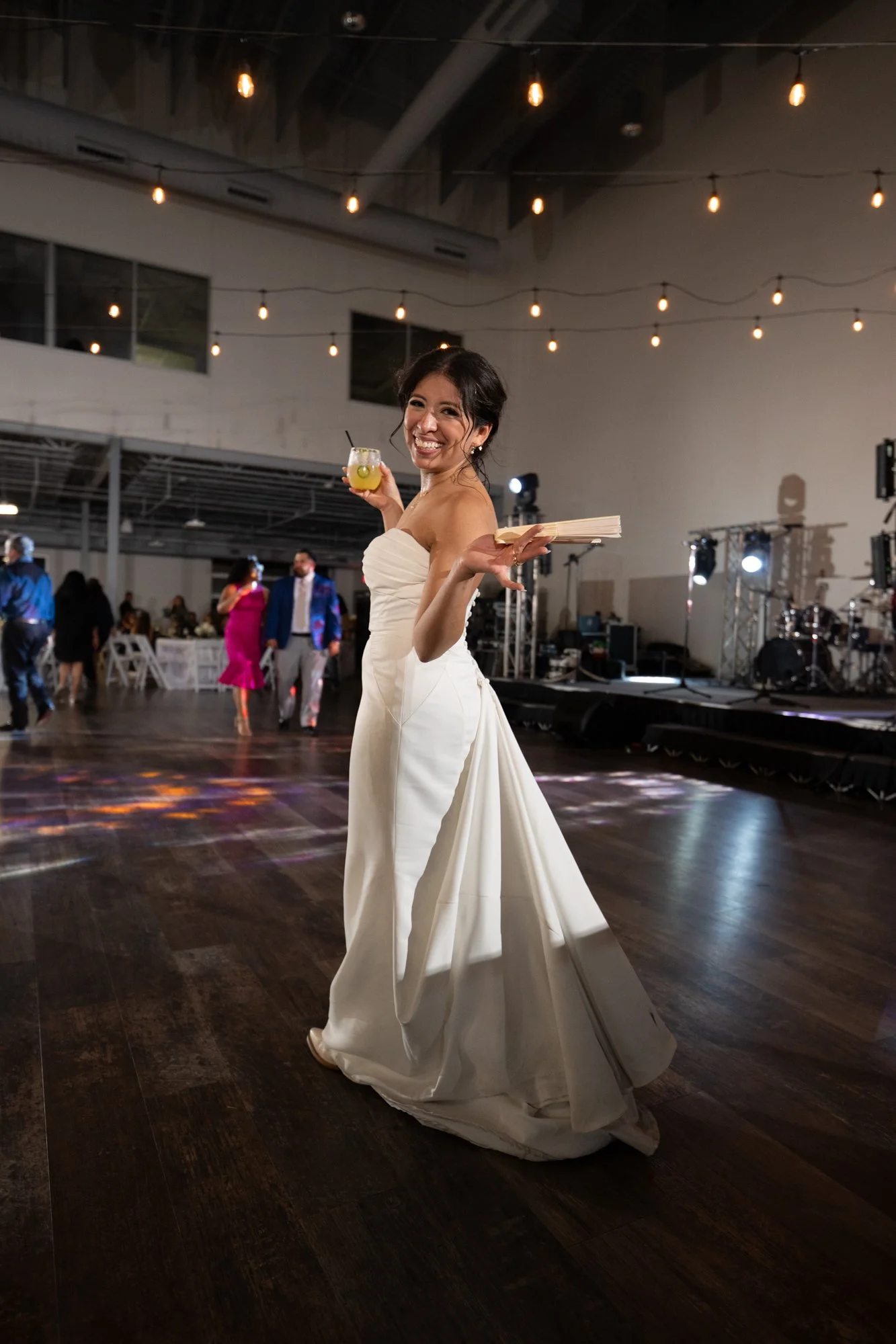 Bride Briseida smiles while walking across the dance floor during her wedding reception in Dallas, Texas, holding a cocktail and celebrating with family and friends.