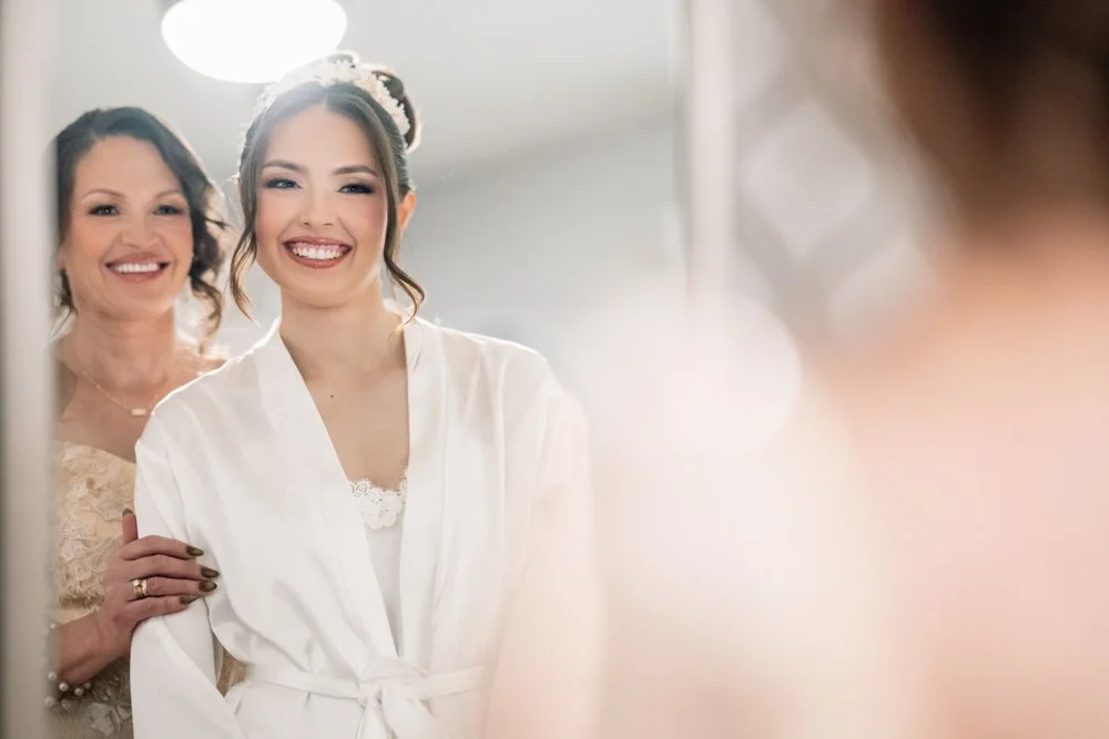 Bride smiling in a bridal suite mirror while getting ready with her mother before the wedding ceremony.