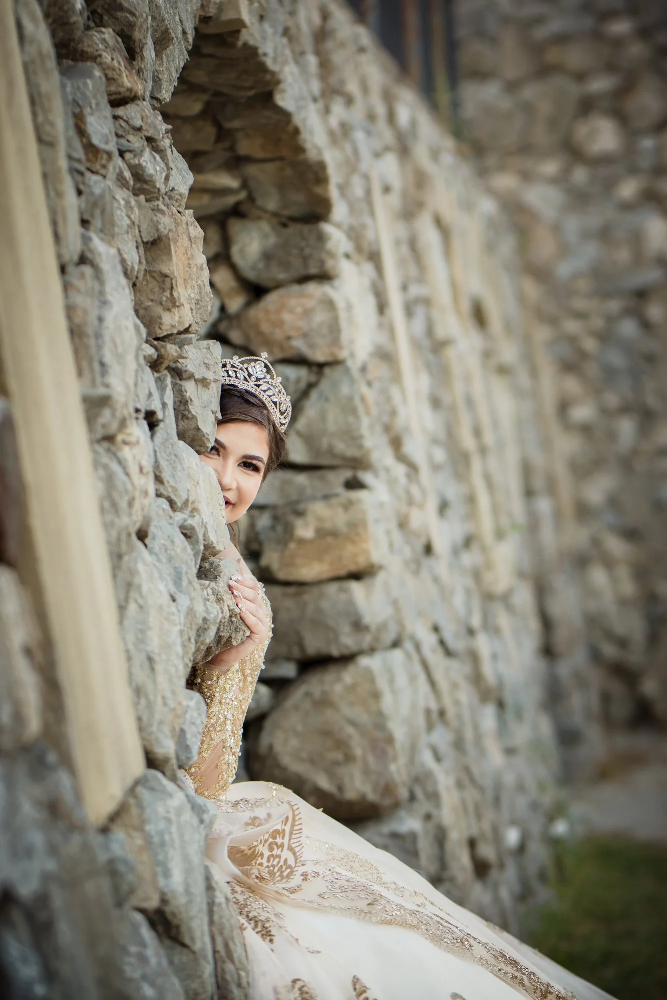 A playful yet elegant moment captured as the Quinceañera peeks from behind a textured stone wall, her crown and detailed gown softly illuminated. This portrait blends personality with sophistication, showcasing confidence, charm, and youthful eleganc