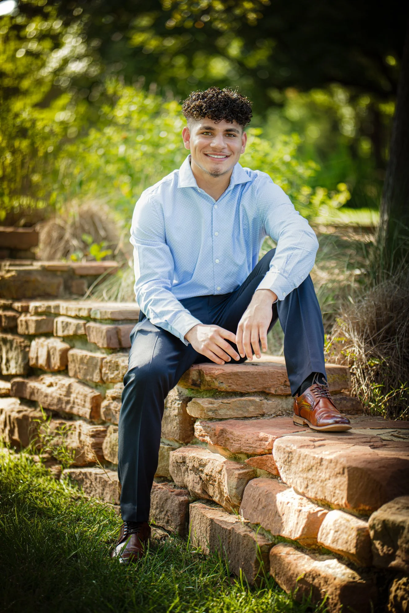A senior portrait of a young man sitting on stone steps outdoors, wearing a light blue dress shirt and navy trousers, smiling confidently in warm natural light.