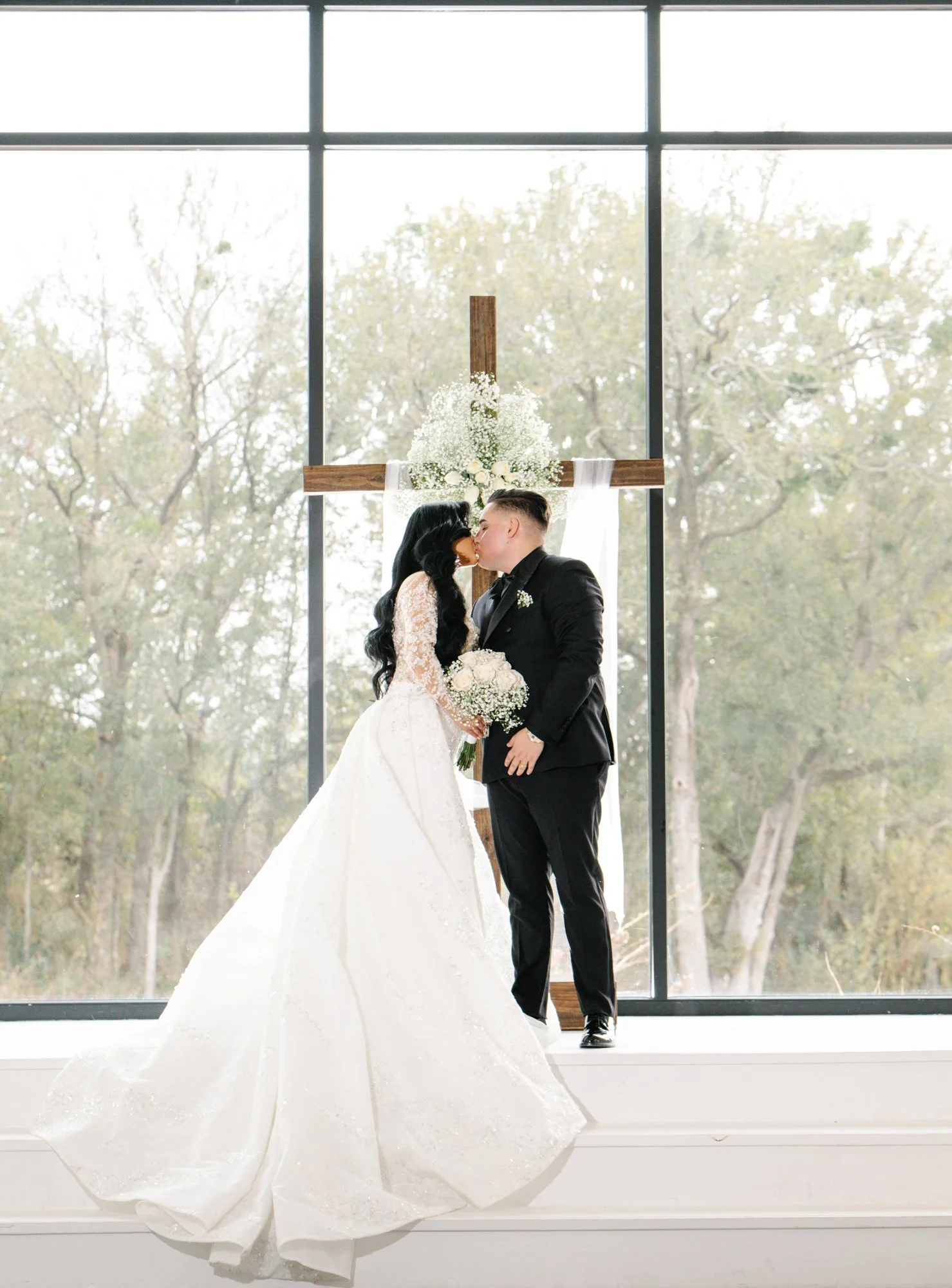 The newlyweds share their first kiss beneath the cross, framed by soft light and floral details, symbolizing faith, unity, and lifelong commitment.