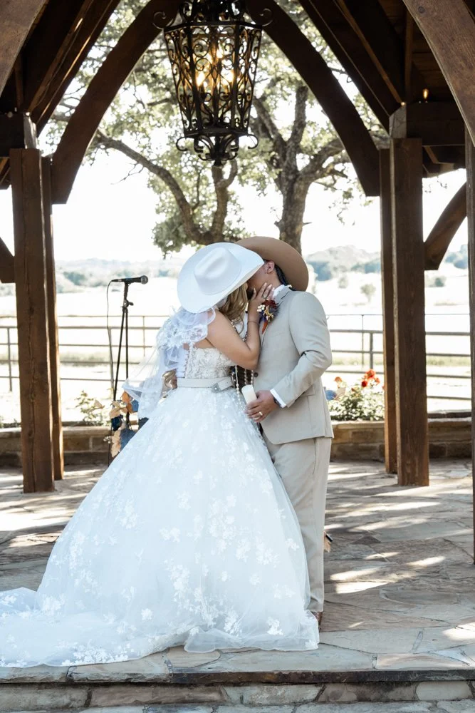 Ashley and Jerman share their first kiss beneath a rustic wooden arbor during their elegant Texas ranch wedding ceremony.