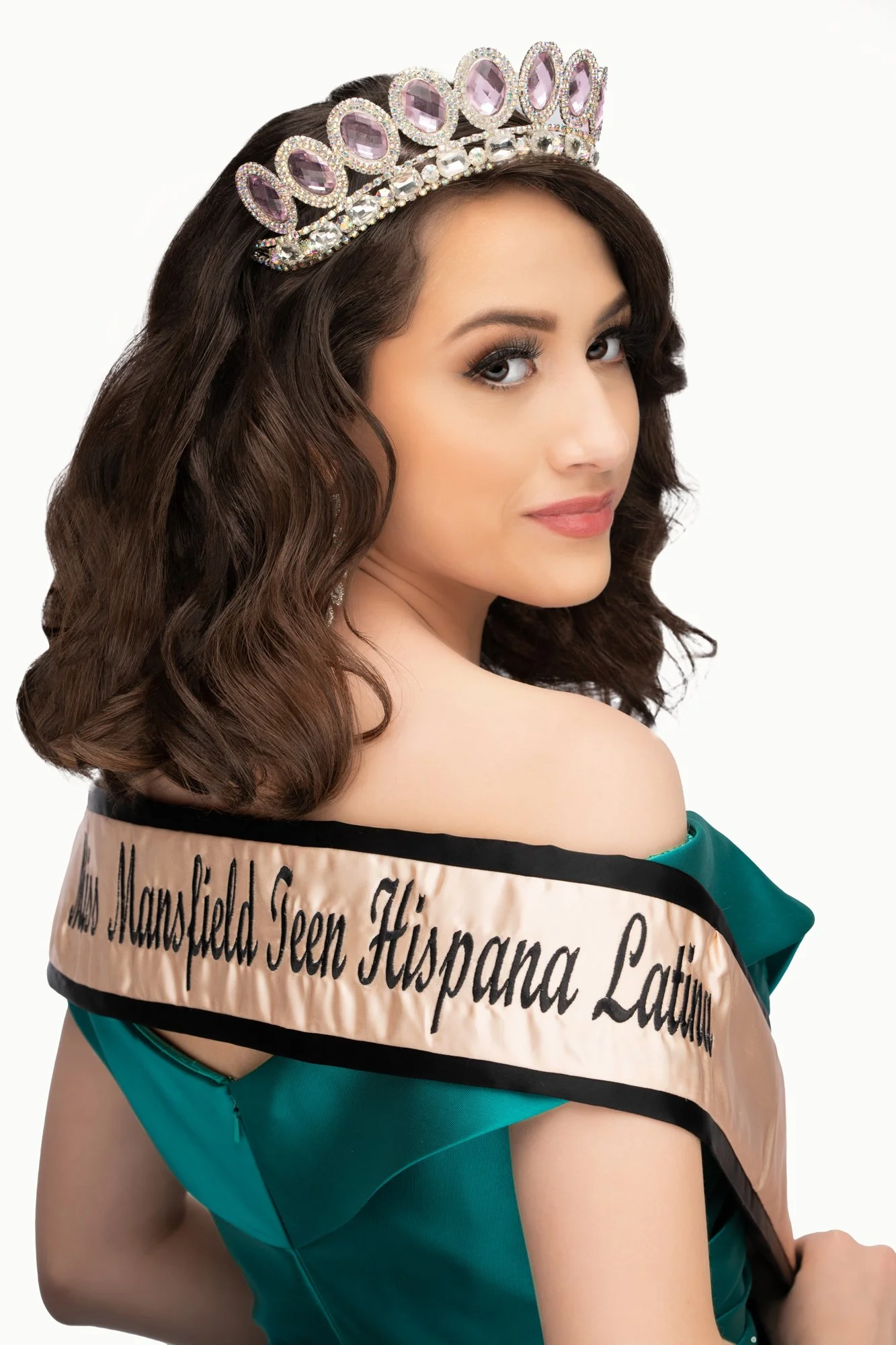A studio portrait of a young woman wearing a jeweled crown and a vibrant emerald green gown, looking over her shoulder while displaying a sash that reads “Mrs. Mansfield Teen Hispana Latina,” captured against a clean white background.