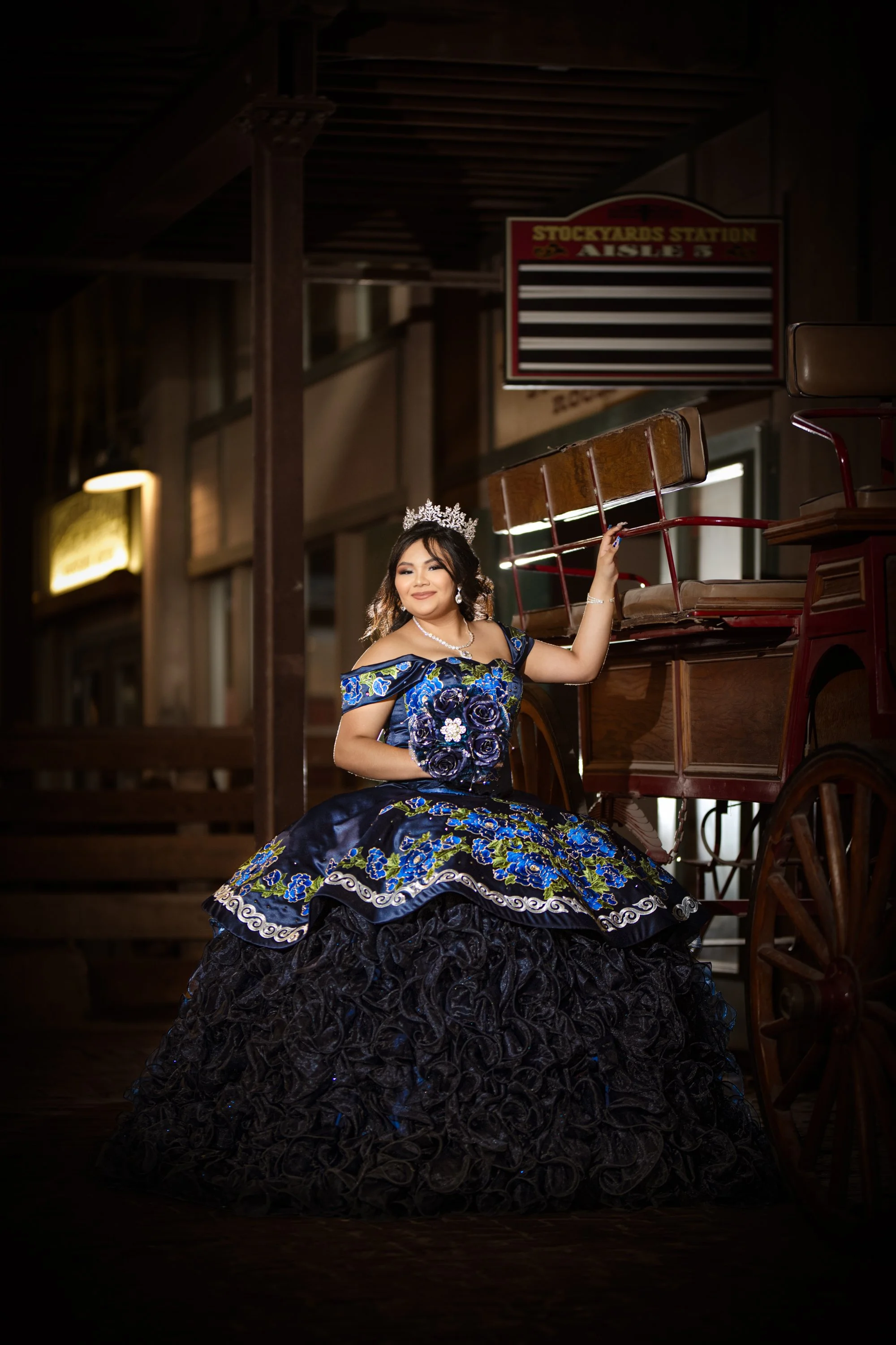 A quinceañera stands gracefully beside a vintage wooden carriage inside a historic stockyards setting, wearing a dramatic navy and royal-blue ball gown with intricate floral embroidery. Her tiara, bouquet, and confident pose create a timeless portrai