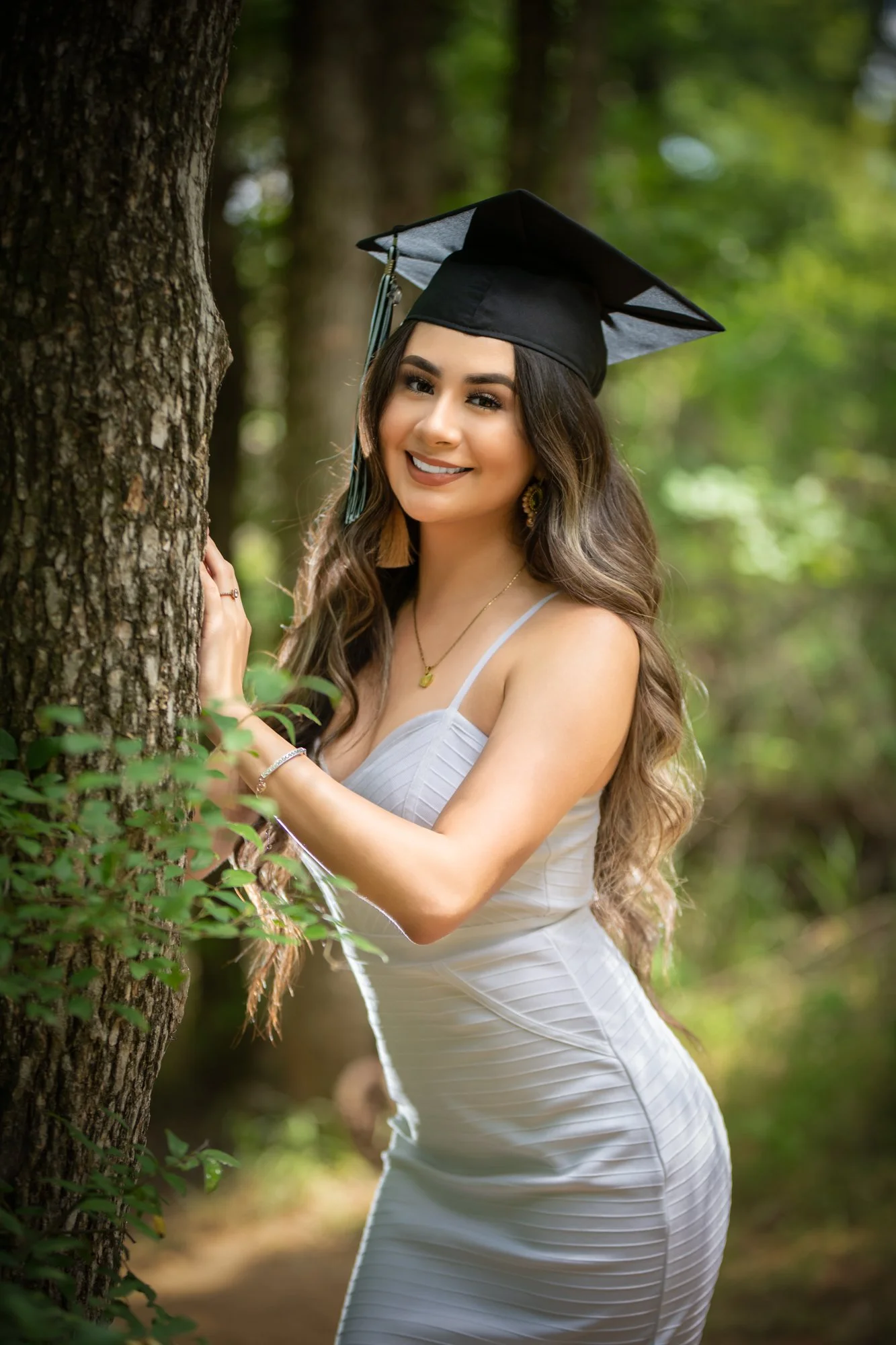A senior portrait of a young woman in a fitted white dress and graduation cap, smiling softly while leaning against a tree in a sunlit wooded park.