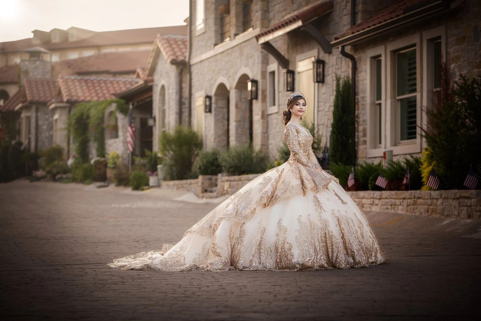 The Quinceañera is photographed walking along a charming stone street lined with historic architecture, her gown trailing behind her in soft light. This image captures movement, tradition, and refined beauty in a cinematic, old-world setting.