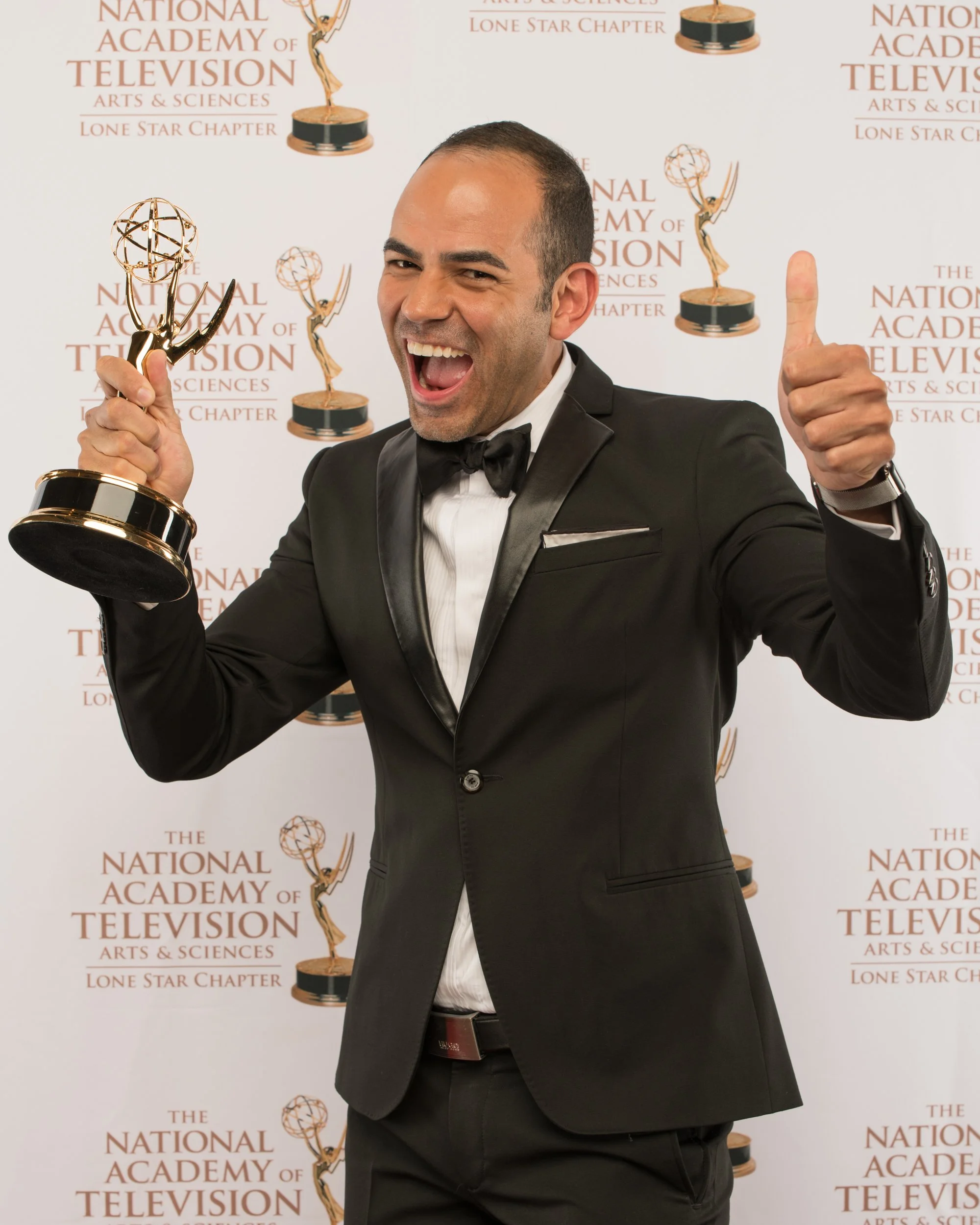 An Emmy Award winner enthusiastically poses with their trophy, giving a thumbs-up while wearing a formal tuxedo in front of the National Academy of Television Arts & Sciences Lone Star Chapter backdrop.